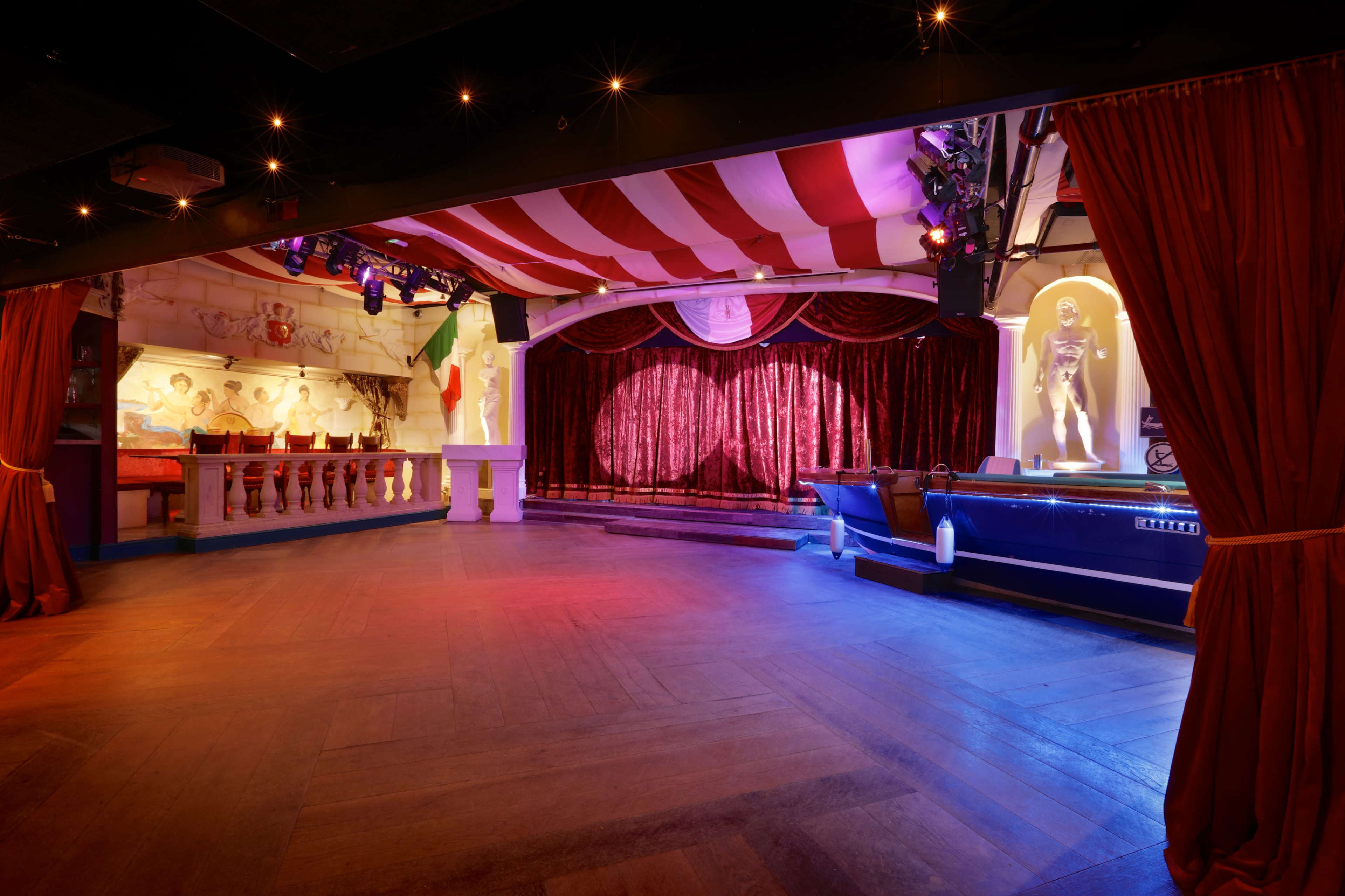 A dimly lit performance space with red velvet curtains, colorful lighting, and a stage featuring decorative elements and a bar area.