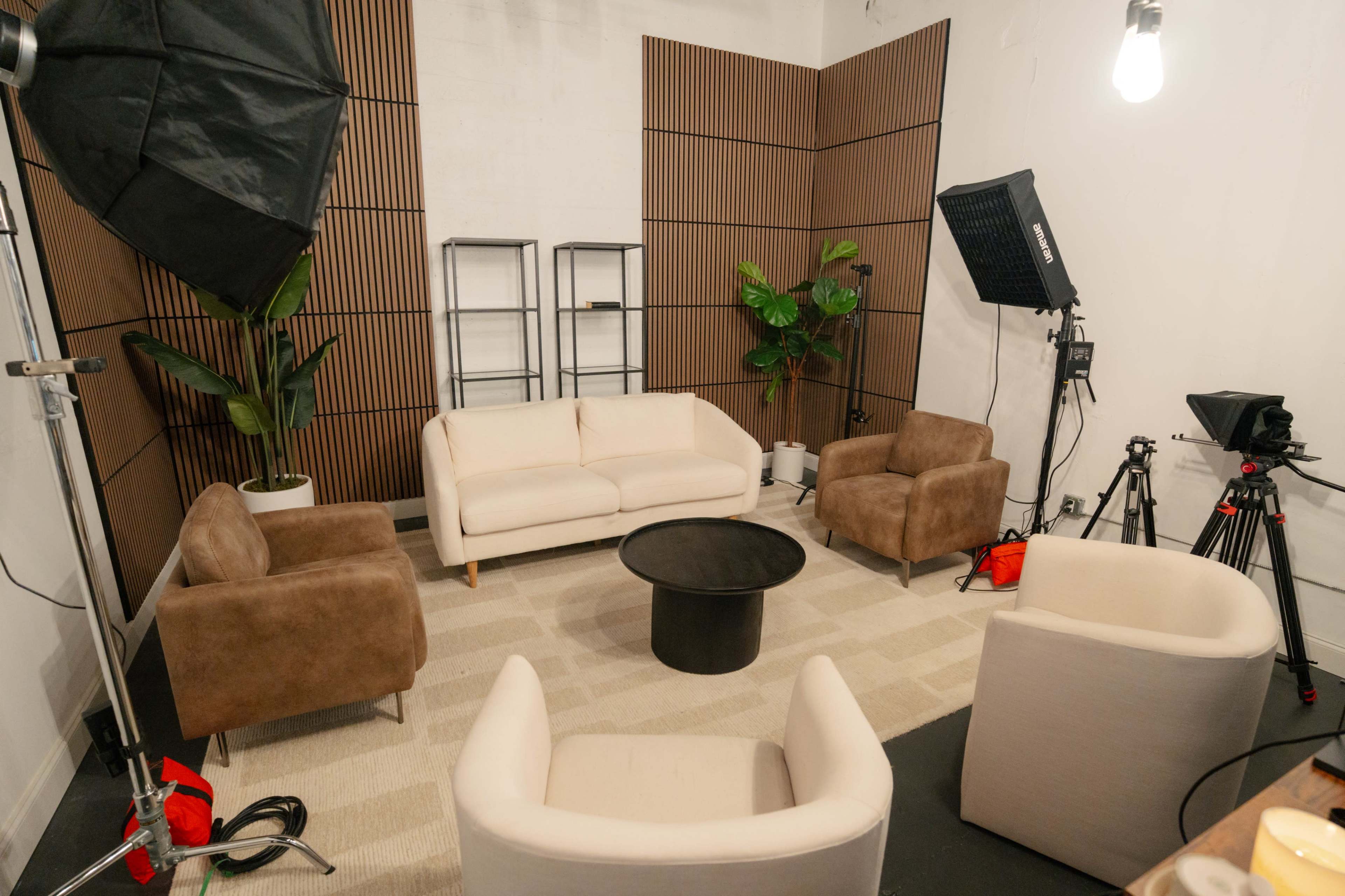 The image shows a studio setup with a light-colored couch, two brown armchairs, and a black table surrounded by various lighting equipment and a plant.
