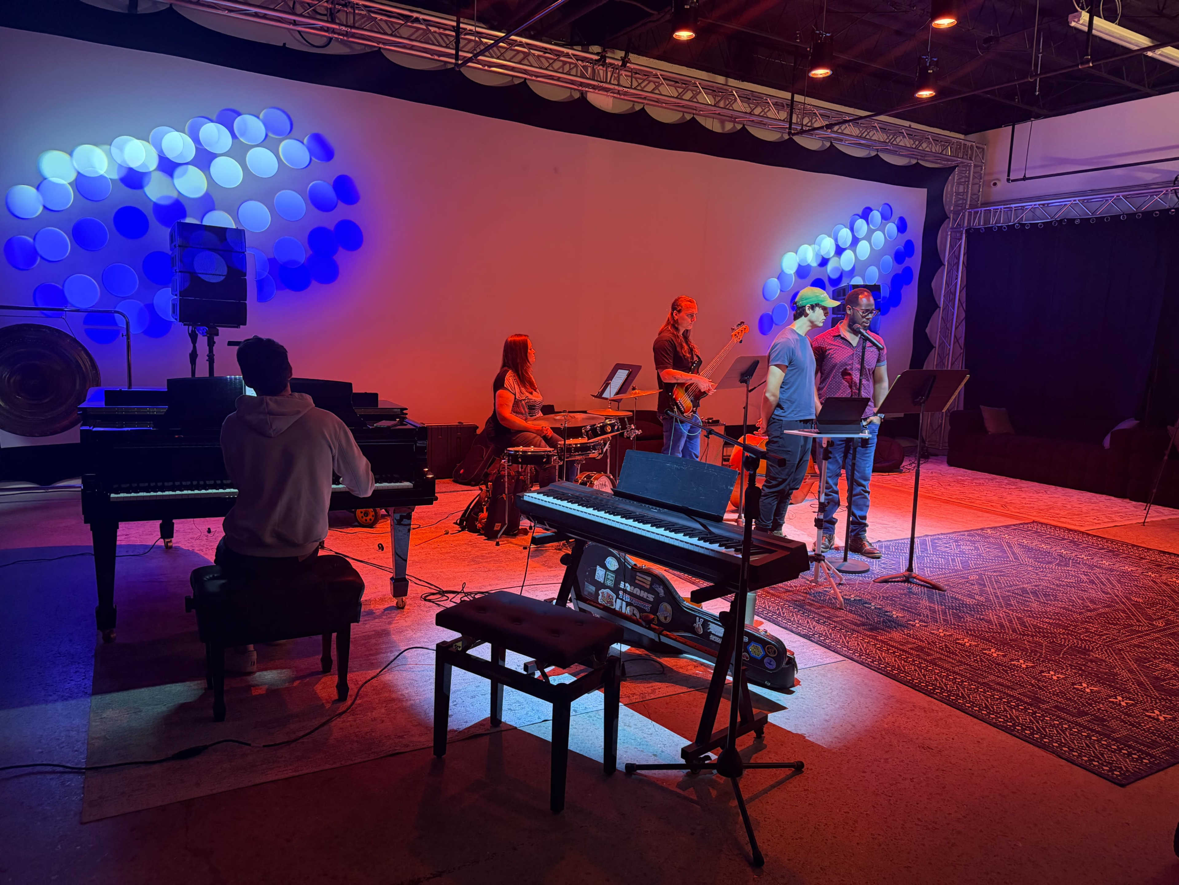A small group of musicians performs on stage, featuring two pianos, drums, and a backdrop with blue and red lighting.
