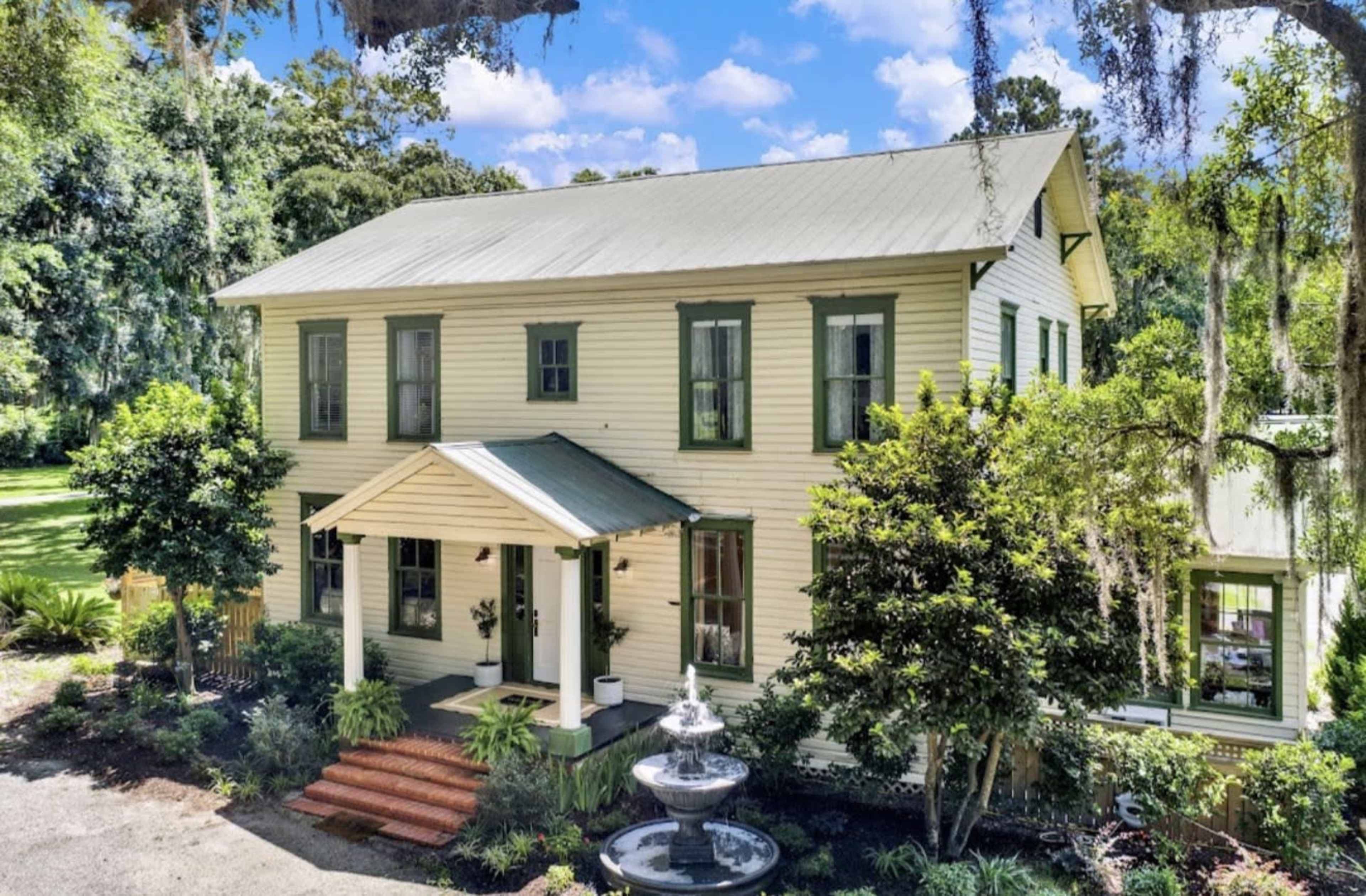 A two-story, light-colored house with green shutters and a front porch is surrounded by lush greenery and a fountain in the front yard.