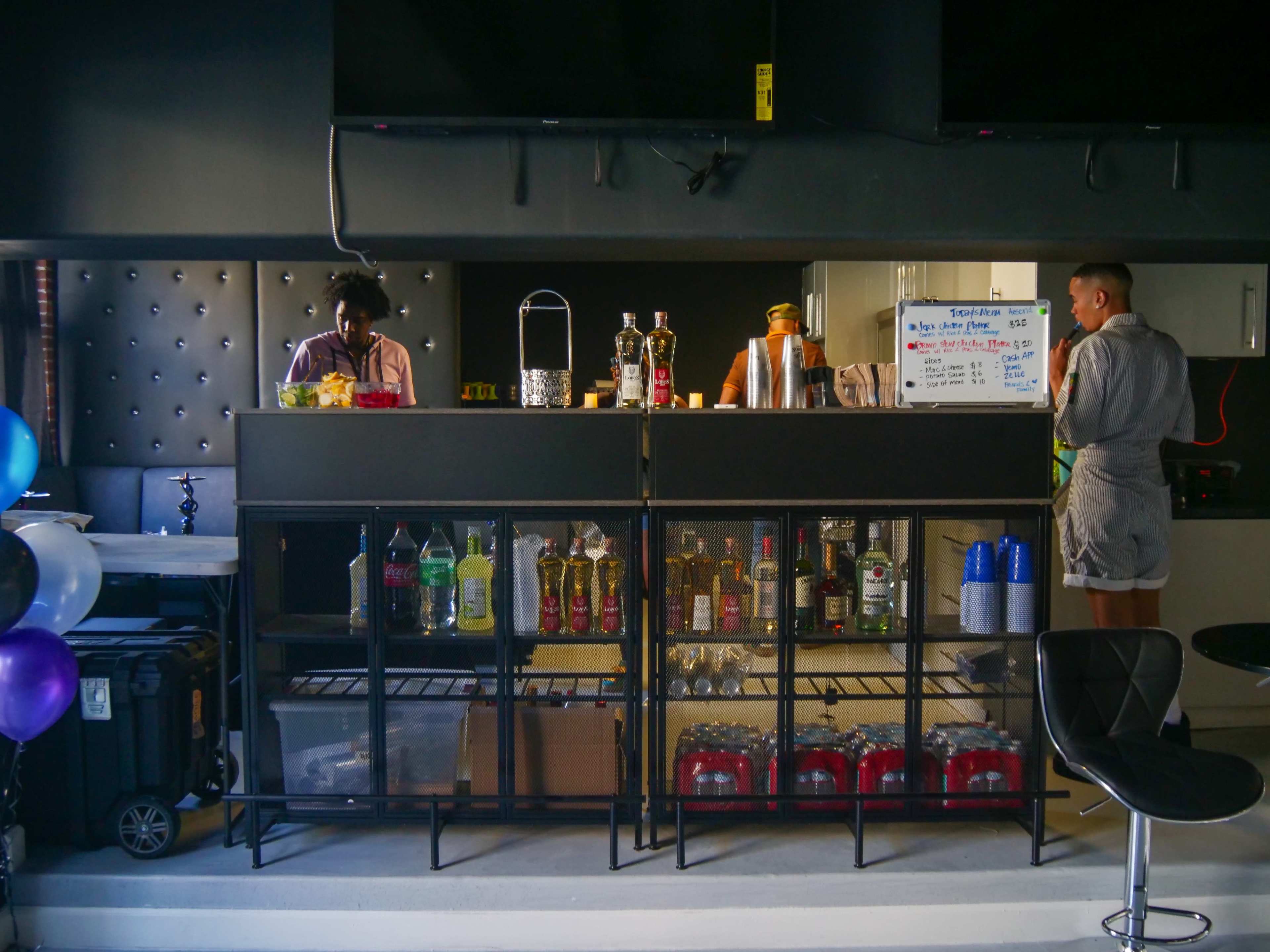 Two individuals prepare drinks at a bar counter filled with various bottles and beverages, while a menu board is visible in the background.