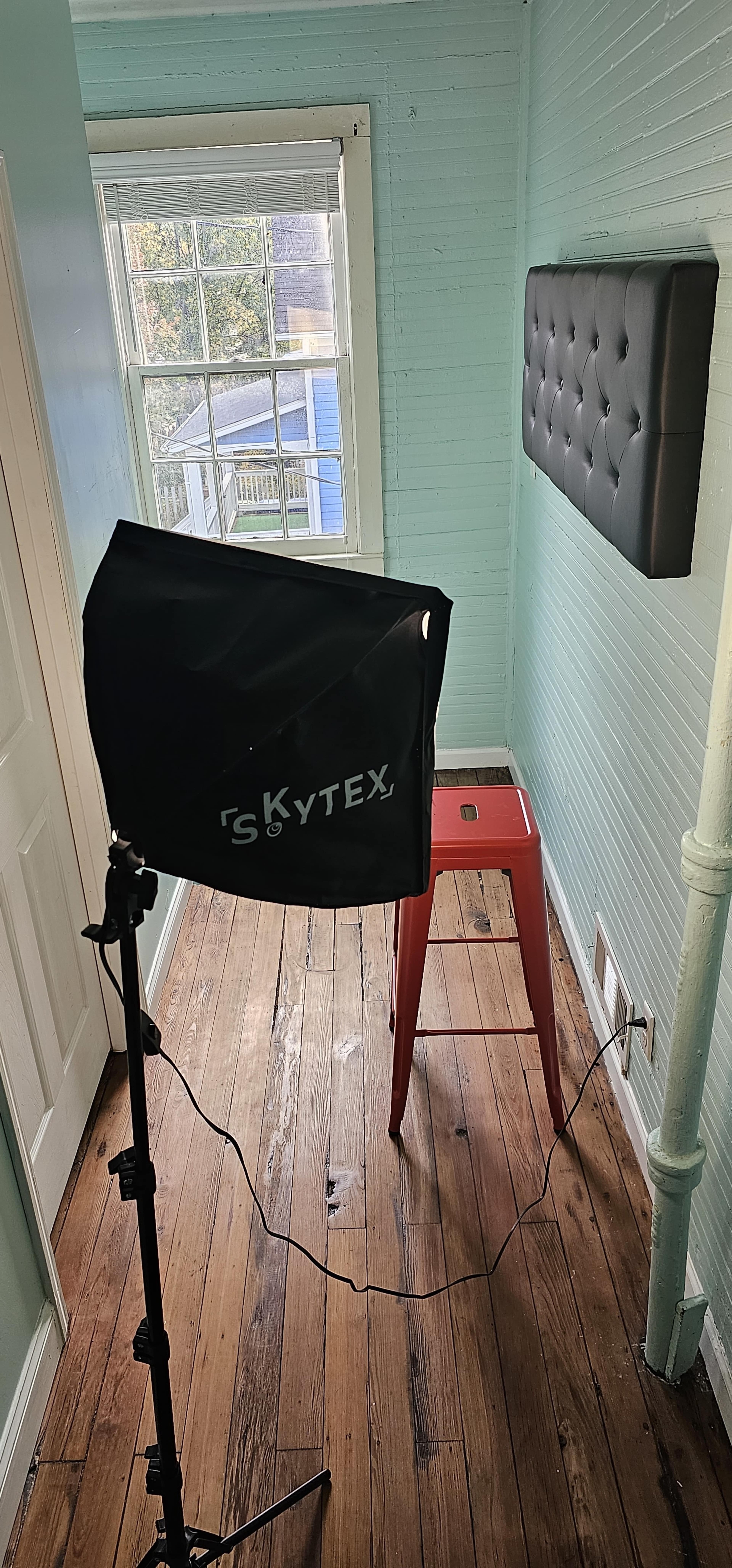 A studio setup features a light umbrella and a red stool positioned in a room with mint green walls and a window.