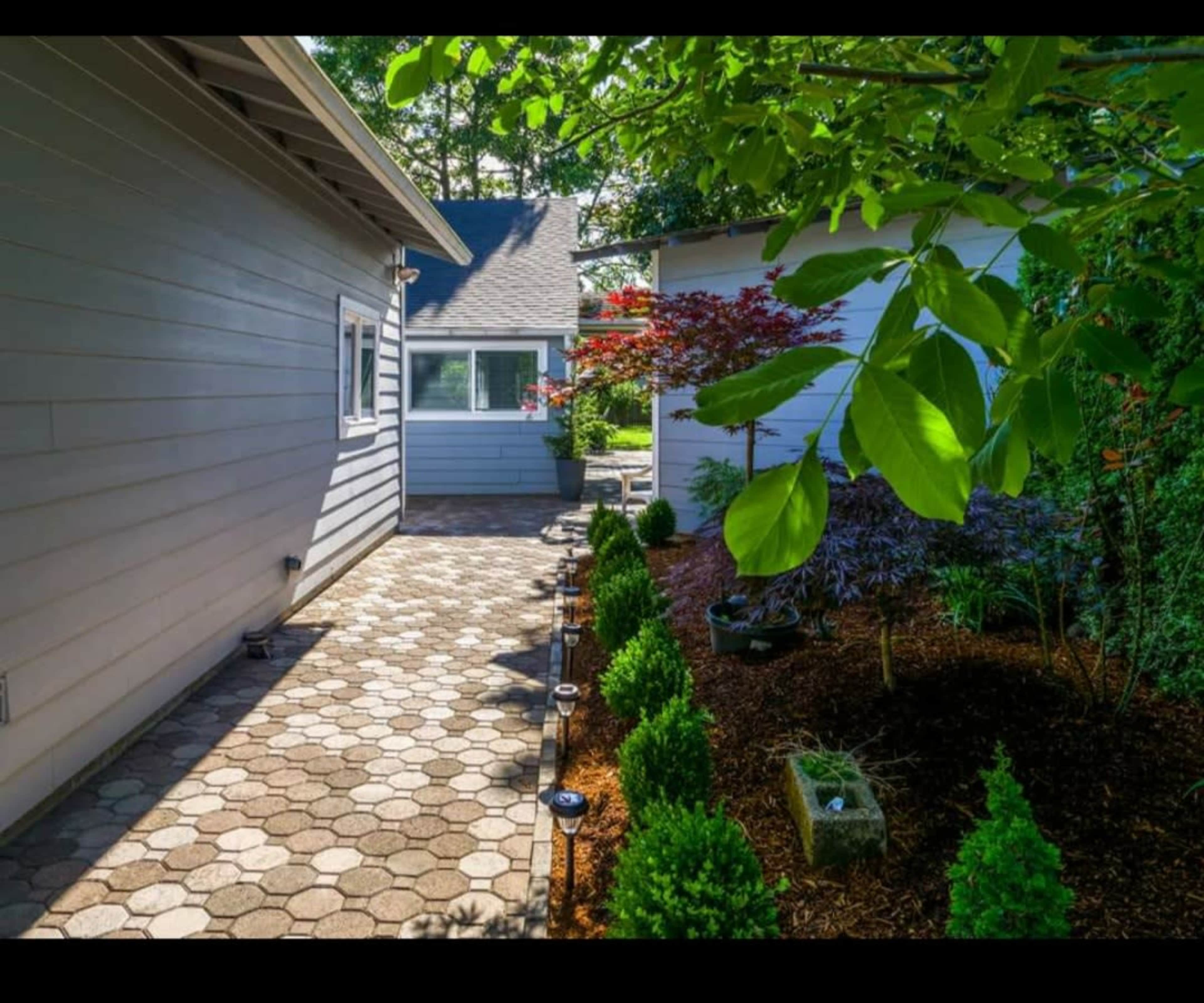 A stone pathway runs between two buildings, bordered by neatly trimmed hedges and landscaped plants under a clear sky.