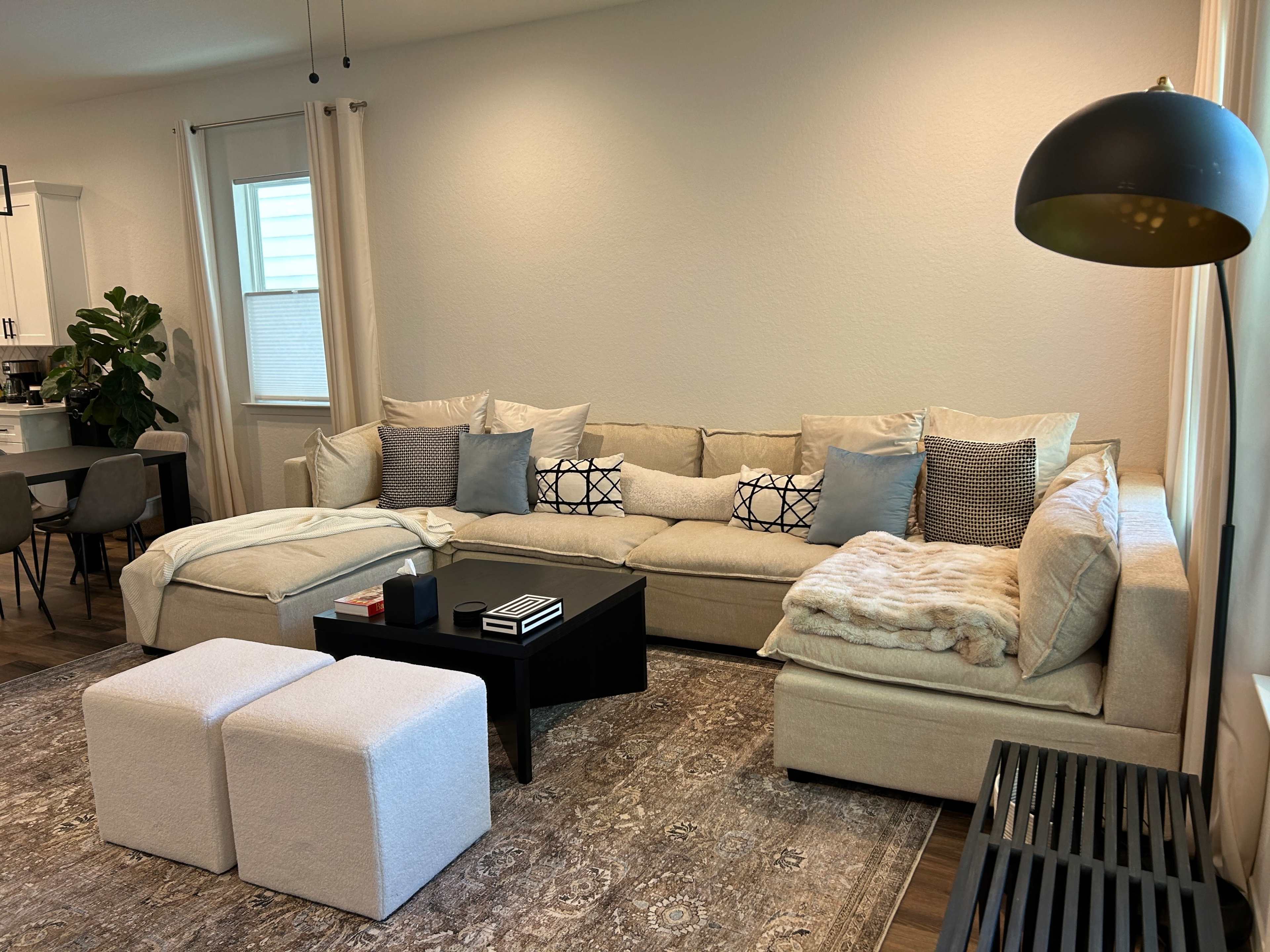 A modern living room with a beige sectional sofa, a black coffee table, and a patterned area rug, along with a dining area in the background.