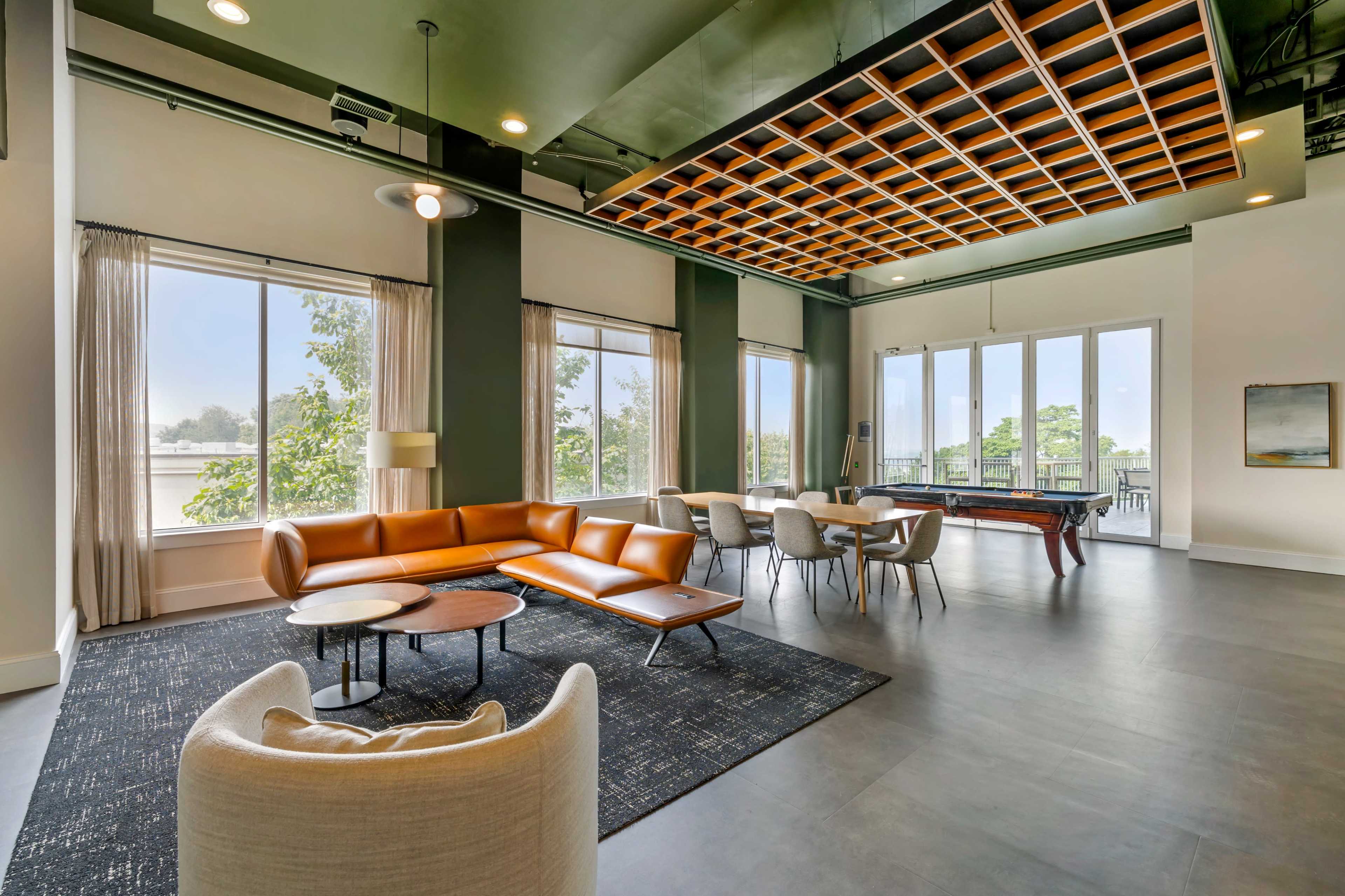 The image shows a modern lounge area featuring large windows, a brown sectional sofa, round tables, and a pool table under a coffered ceiling.