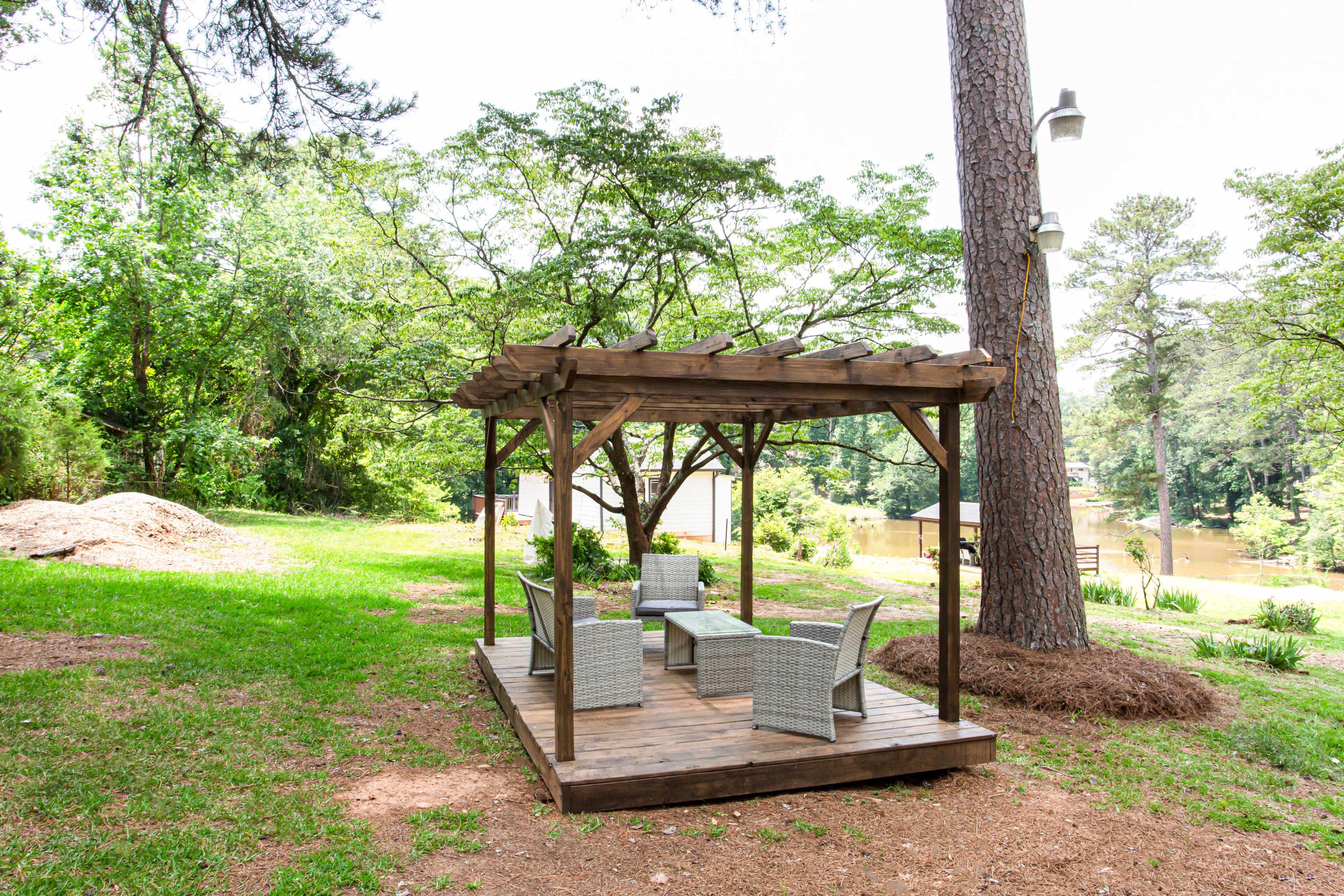 A wooden gazebo with four chairs and a table is set on a grassy area near a river, surrounded by trees.