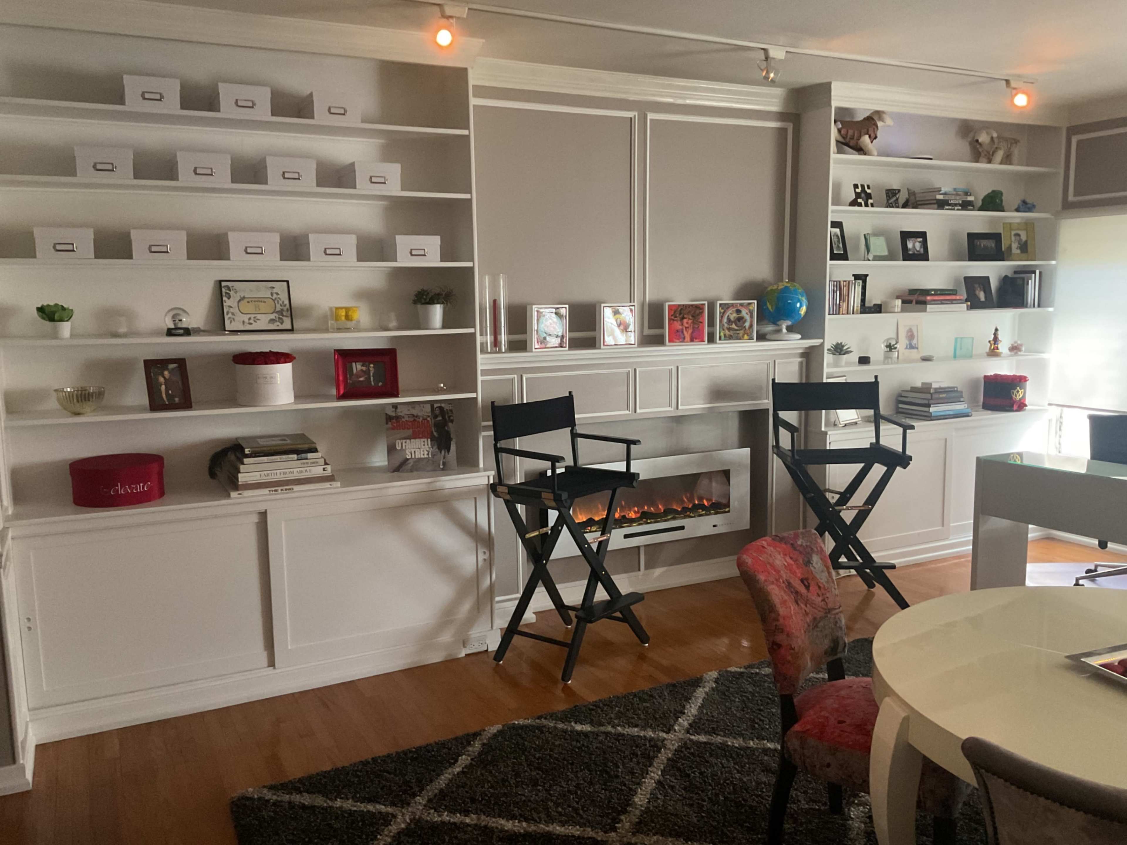 A modern living room features built-in shelves displaying decorative items and books, two director's chairs, and a faux fireplace.