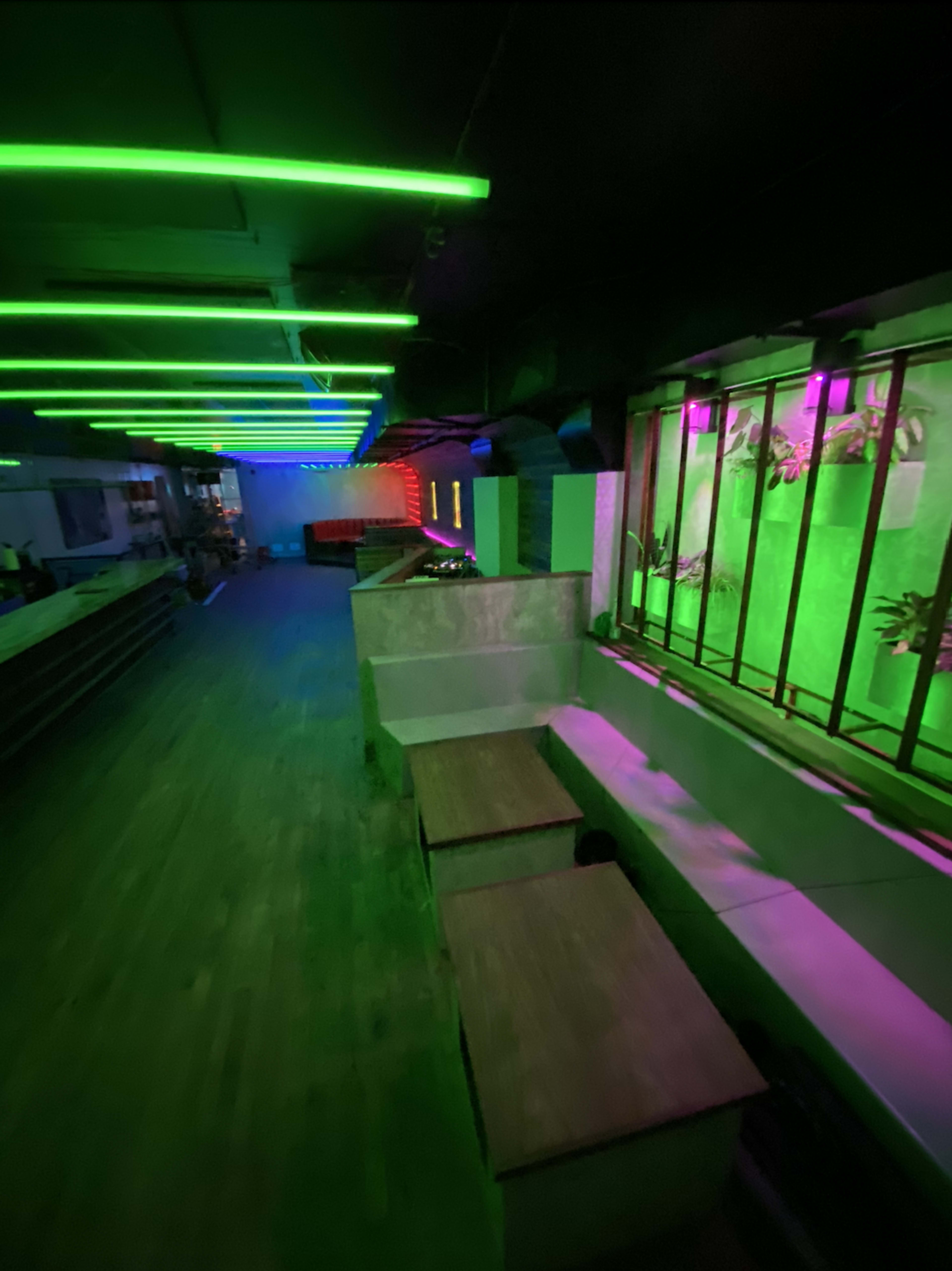 The image shows an empty interior space with benches and glowing neon lights in green and pink.