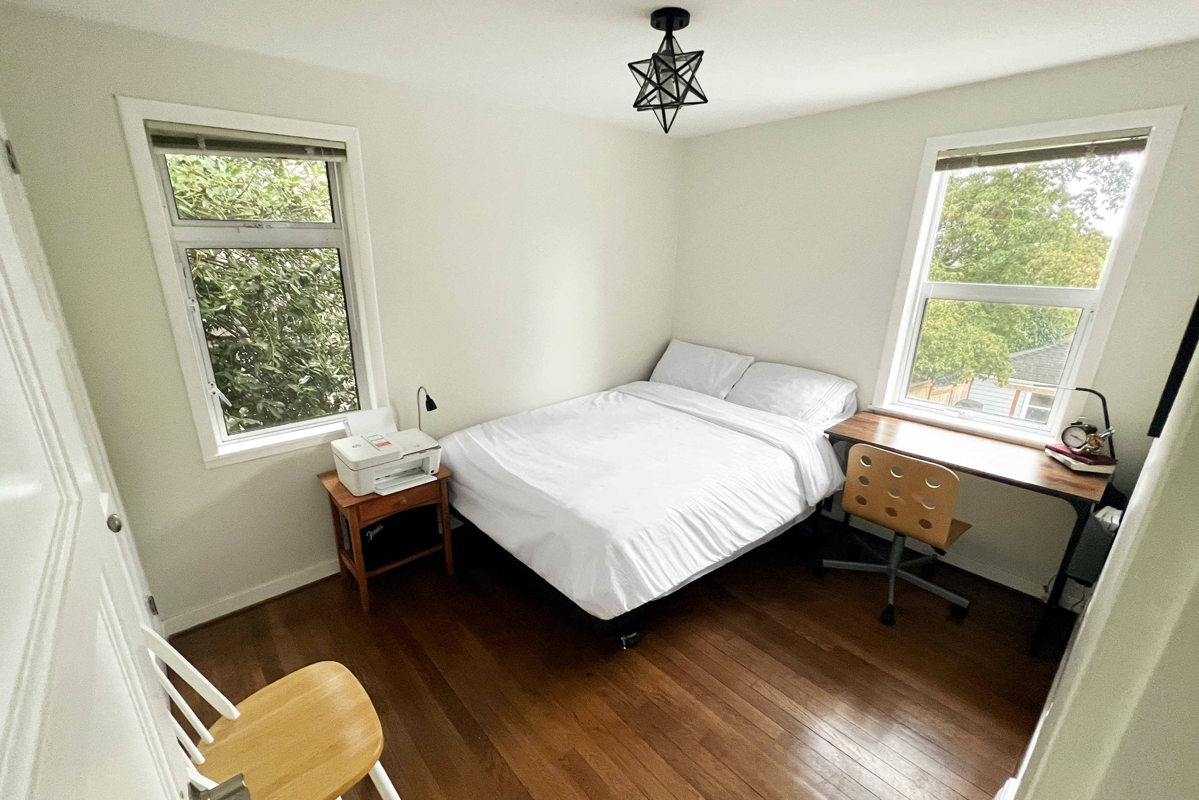 A bright bedroom features a bed with white linens, a wooden desk with a chair, and two windows providing natural light.
