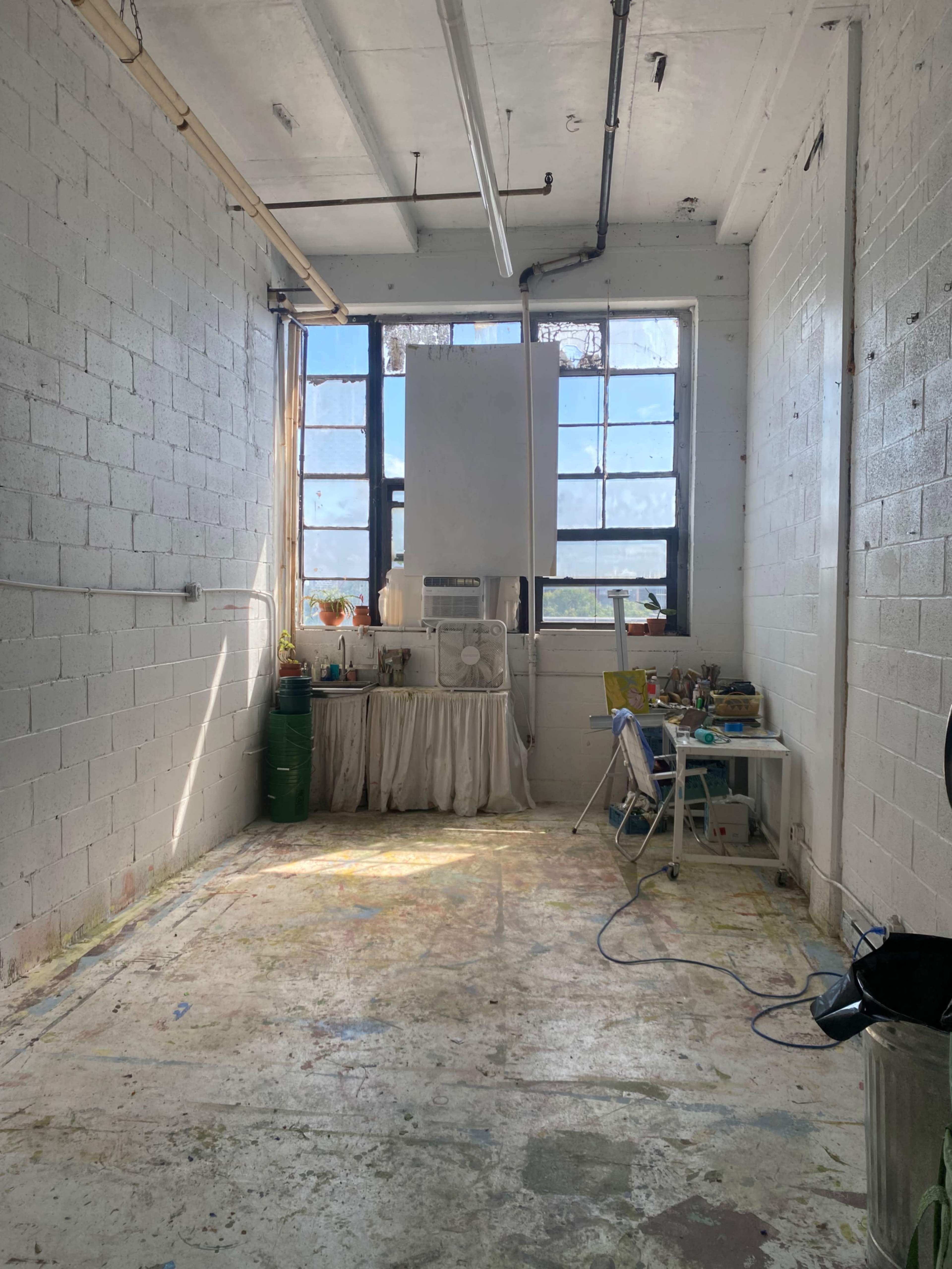 The image shows a spacious, empty artist's studio with large windows, a small table and chair, and scattered art supplies on the floor.