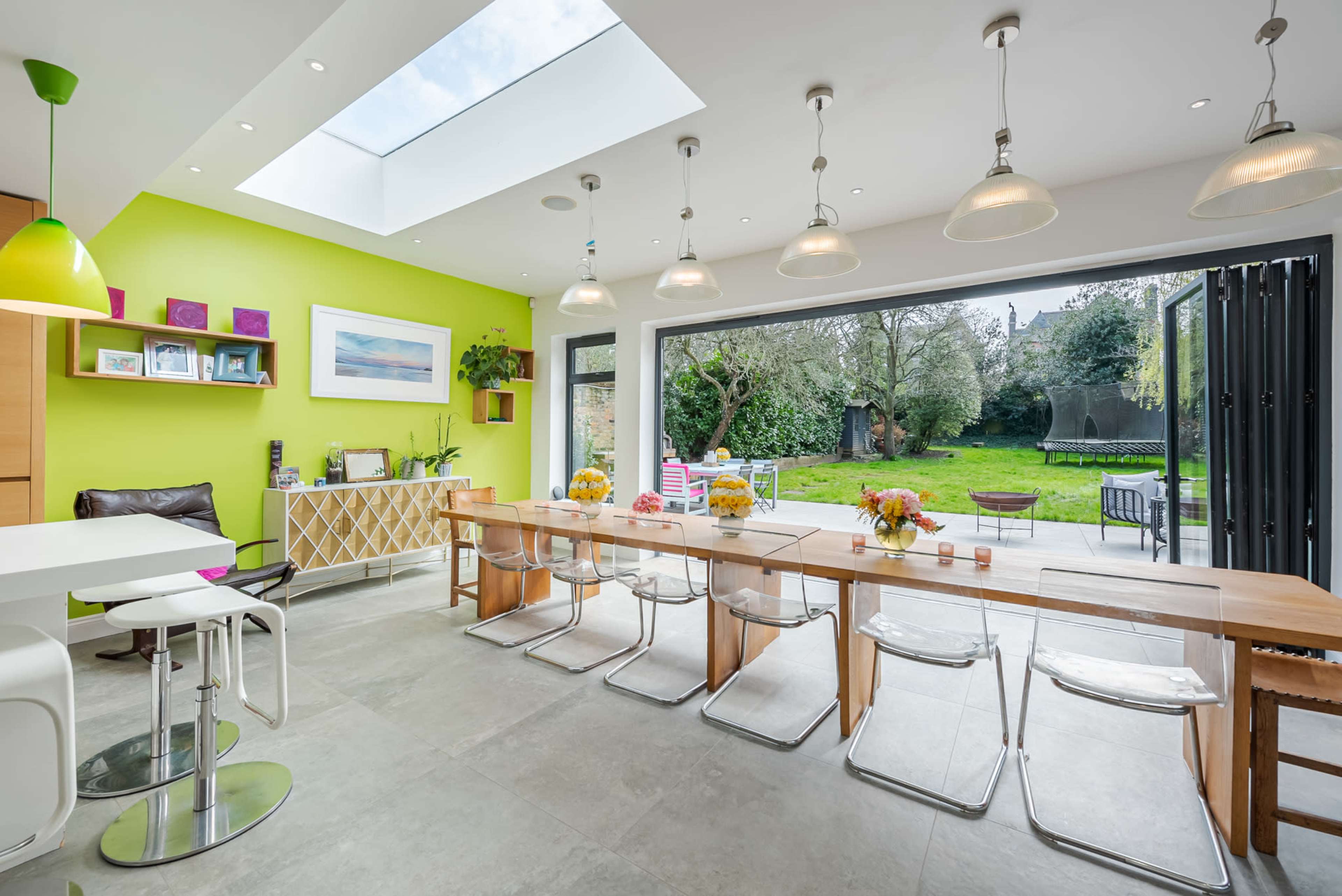 A modern kitchen features a long wooden dining table with clear chairs, large glass doors opening to a garden, and a bright green accent wall.