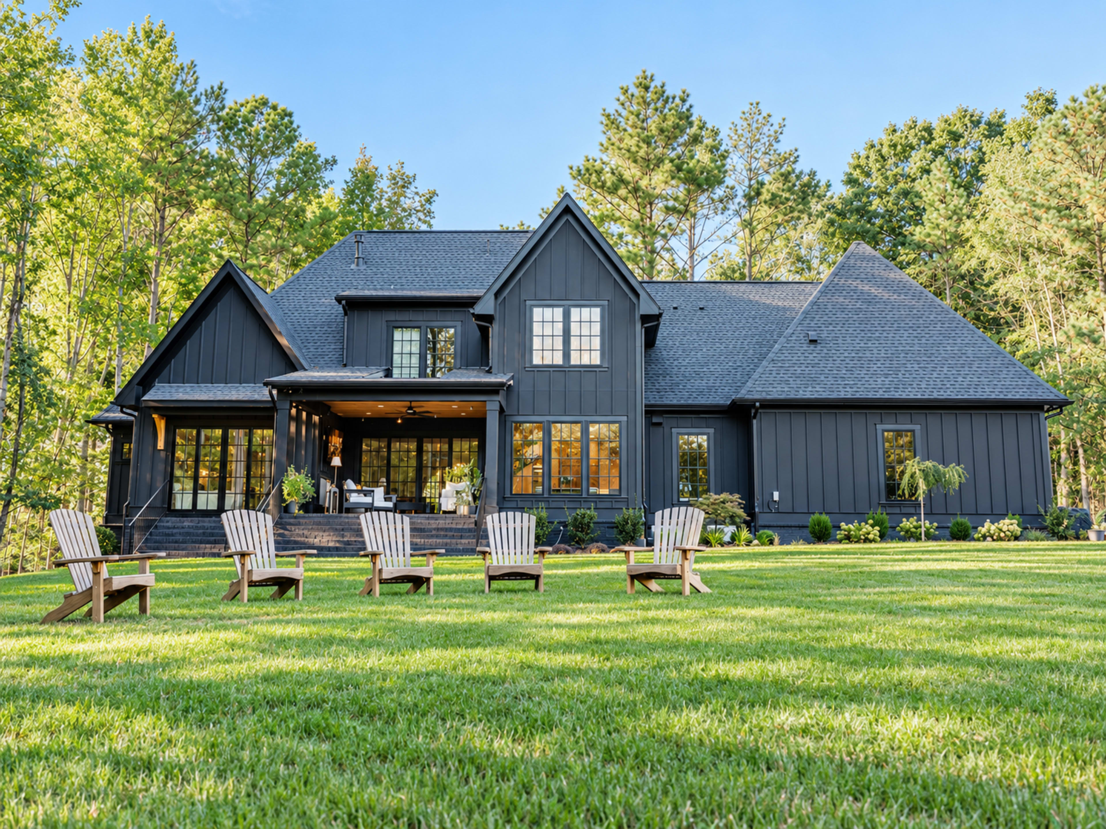 A large, modern black house is surrounded by trees, with a spacious lawn featuring several wooden Adirondack chairs arranged in a row.