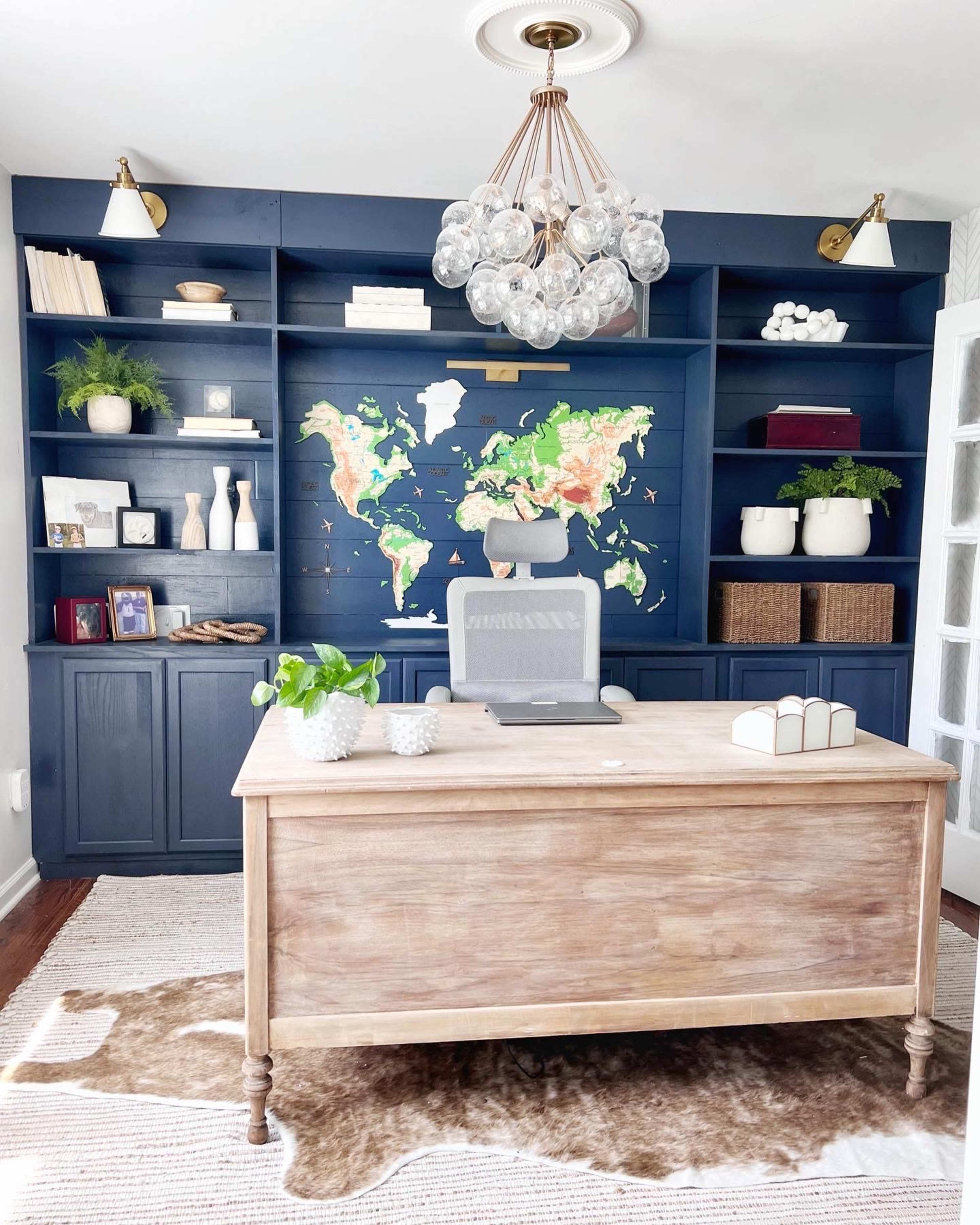 A modern office features a wooden desk facing a wall-mounted world map, with shelves displaying decorative items and plants in a blue-paneled room.