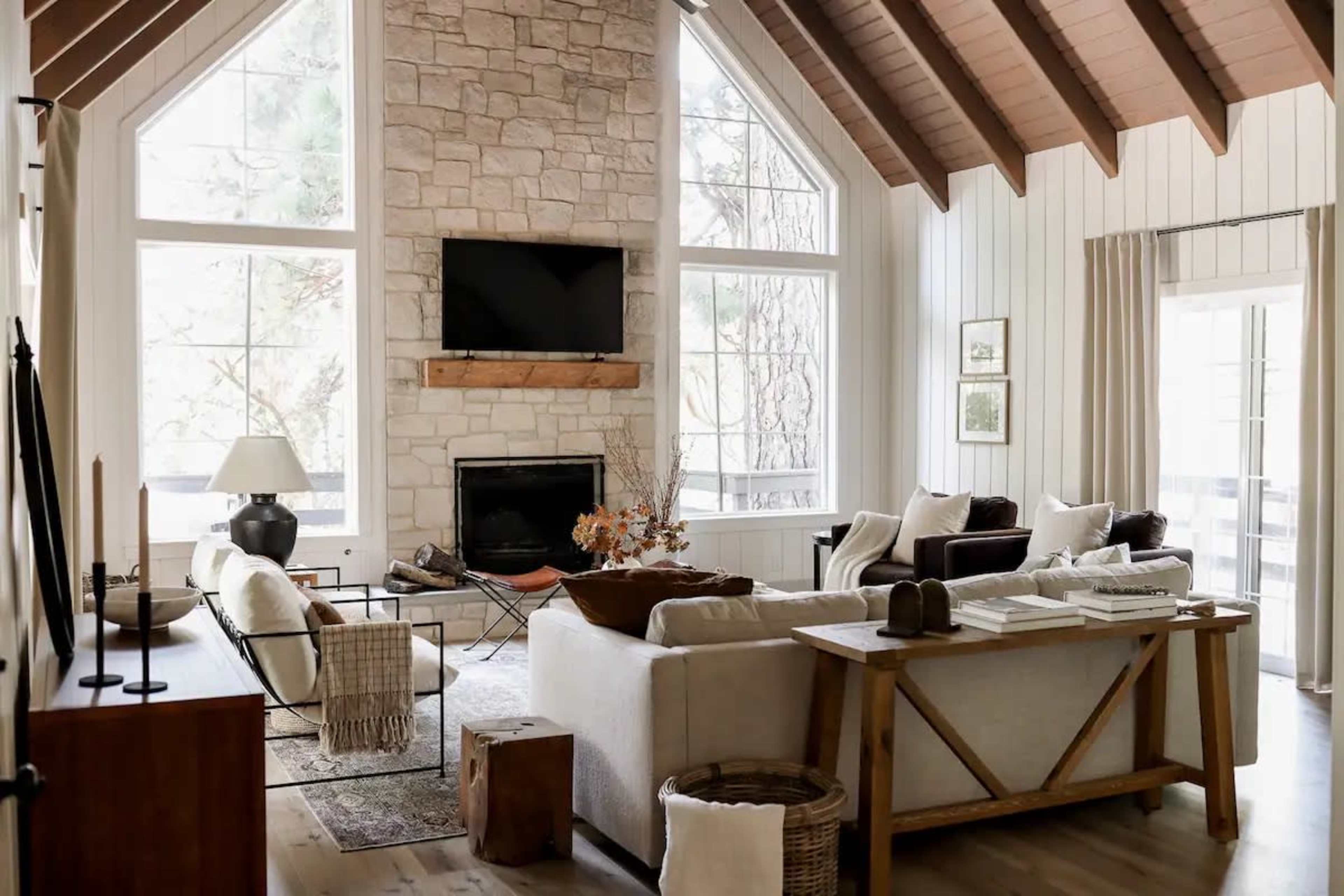 The image shows a bright living room featuring a stone fireplace, large windows, and a mix of modern and rustic furniture arrangements.