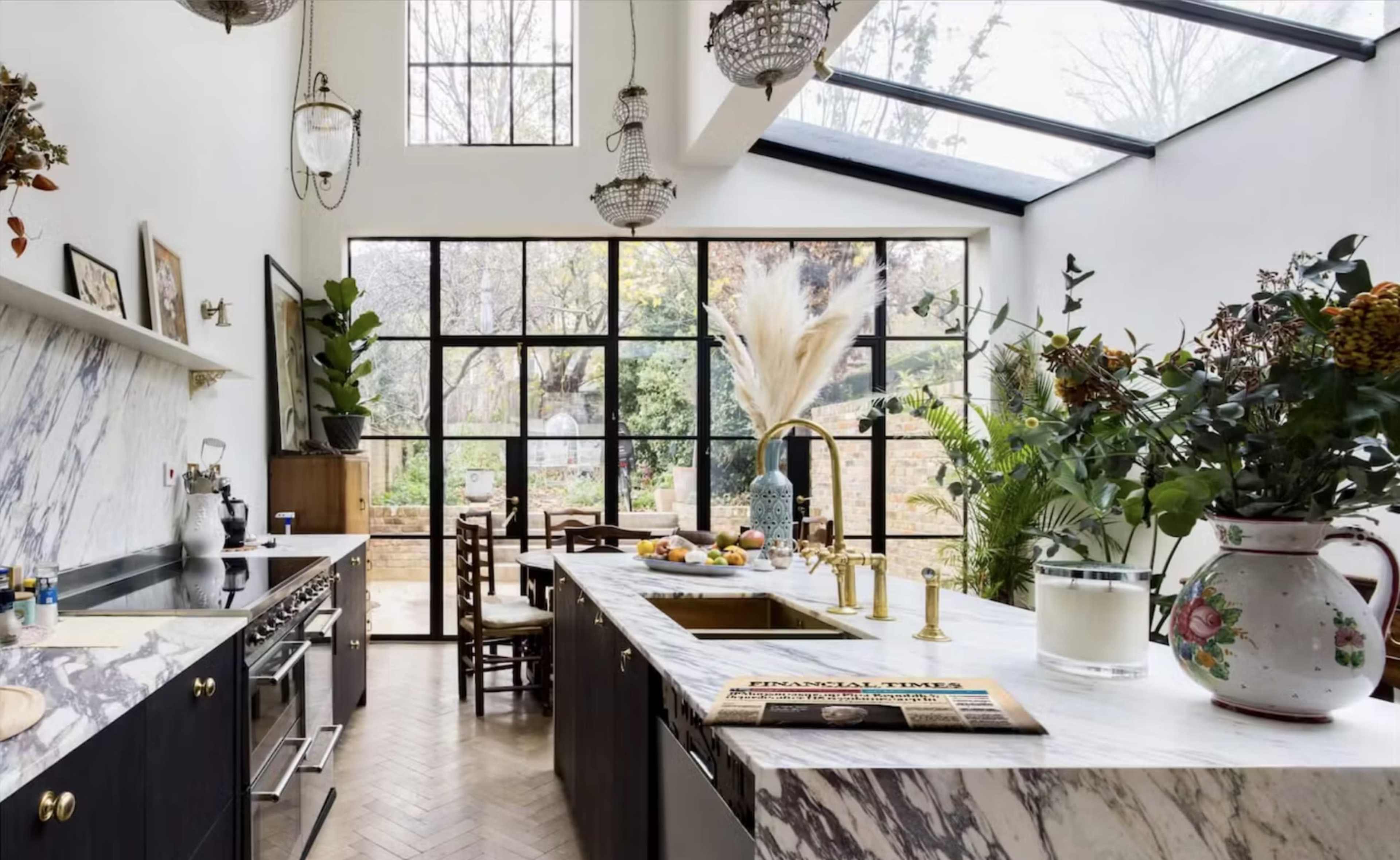 The kitchen features a large marble island, glass walls, and an abundance of plants and natural light.