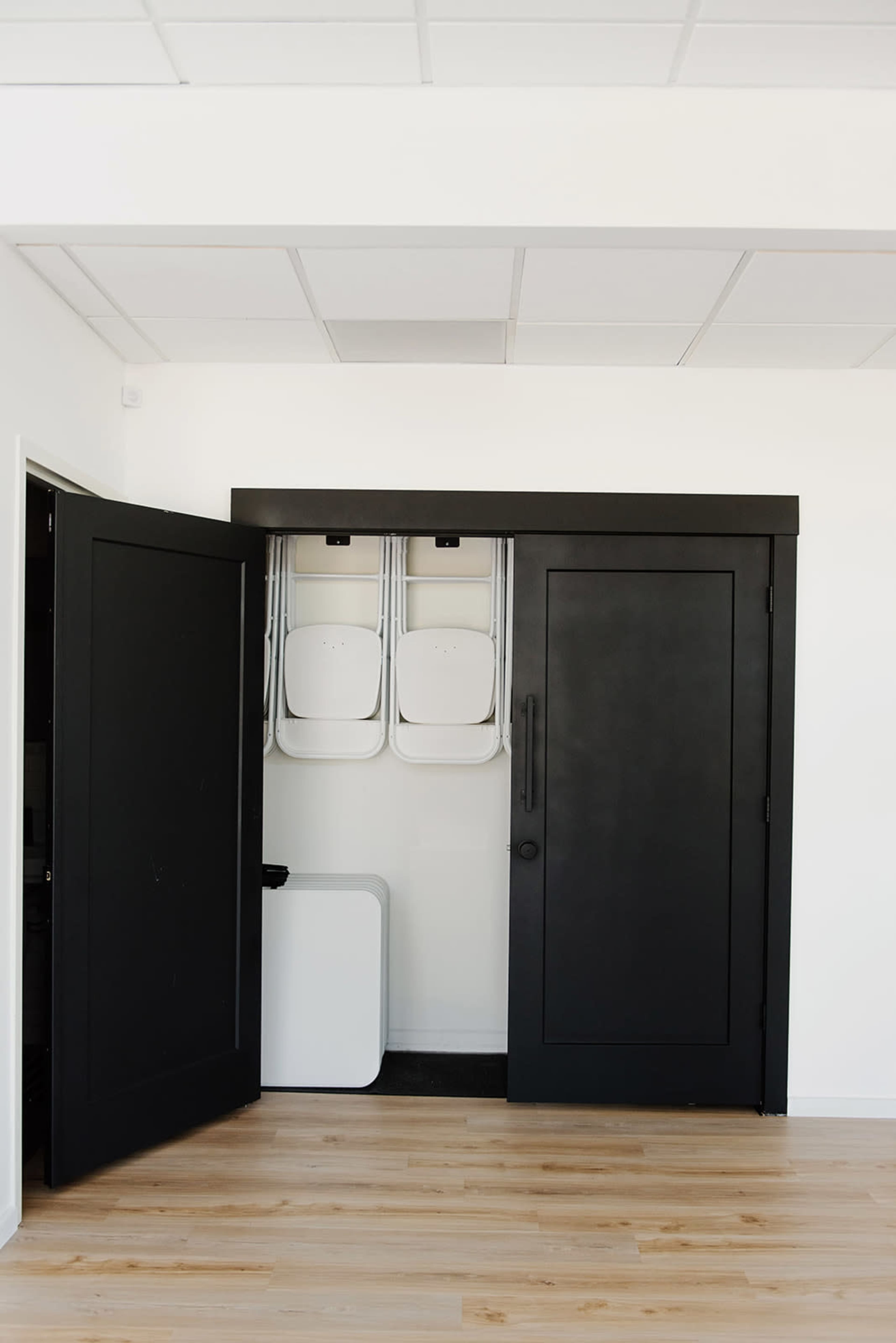 The image shows an open black double door revealing a storage area with stacked folding chairs and a white object.