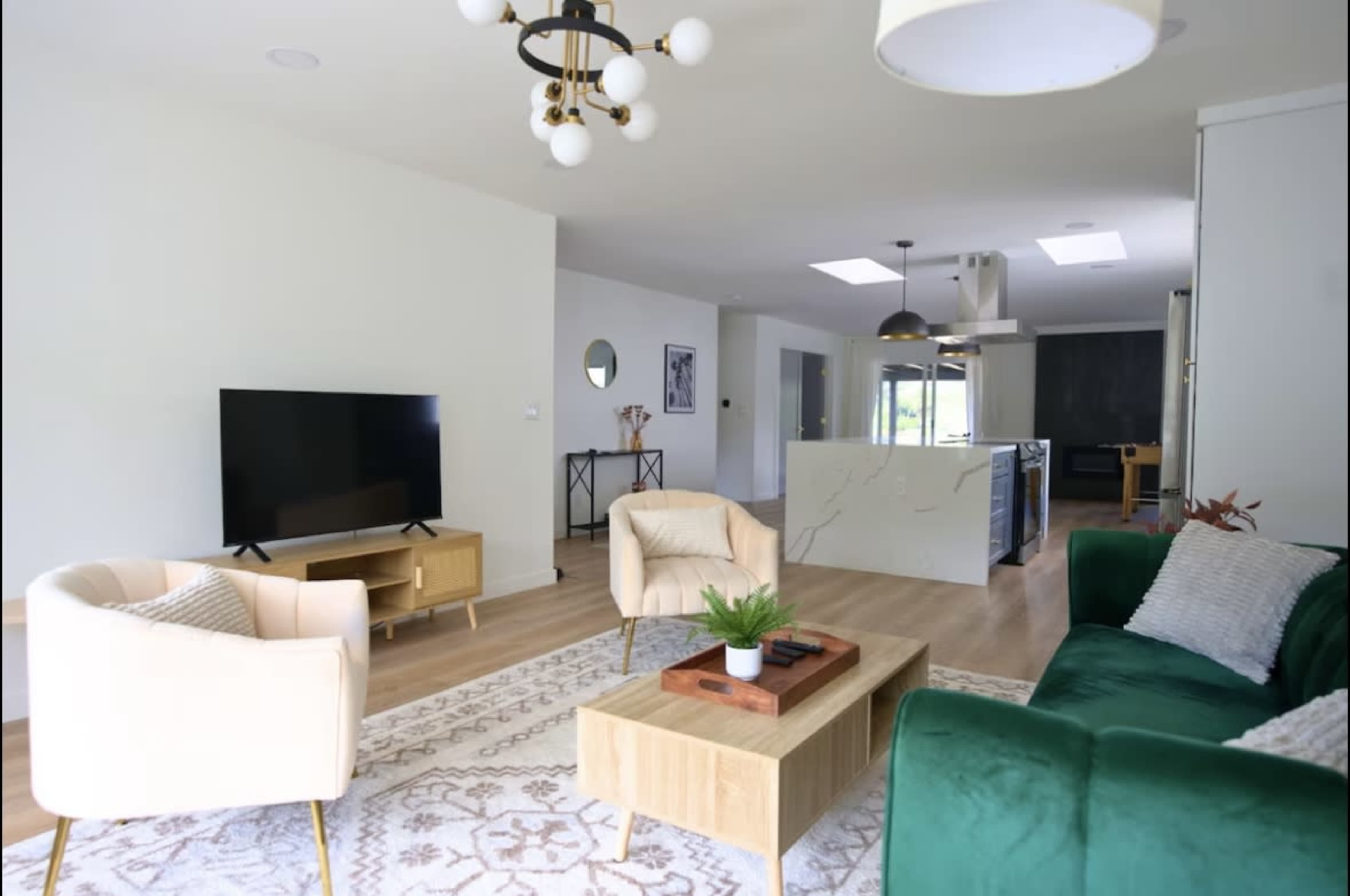 The image shows a modern living room with two armchairs, a green sofa, a wooden coffee table, a television on a stand, and an open kitchen area in the background.