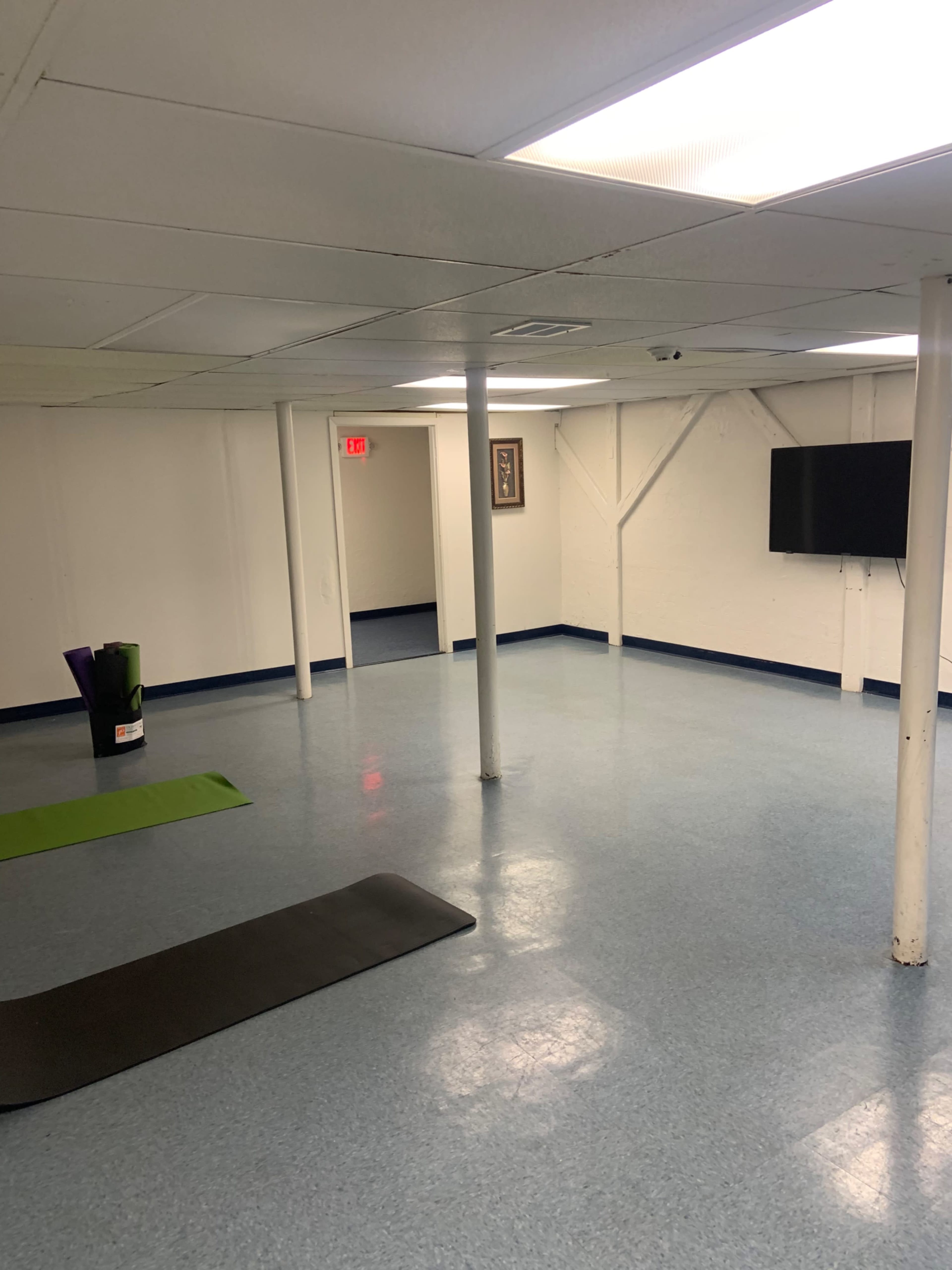 A spacious, empty basement room with blue flooring, a yoga mat, a green exercise mat, a television mounted on the wall, and structural support poles.