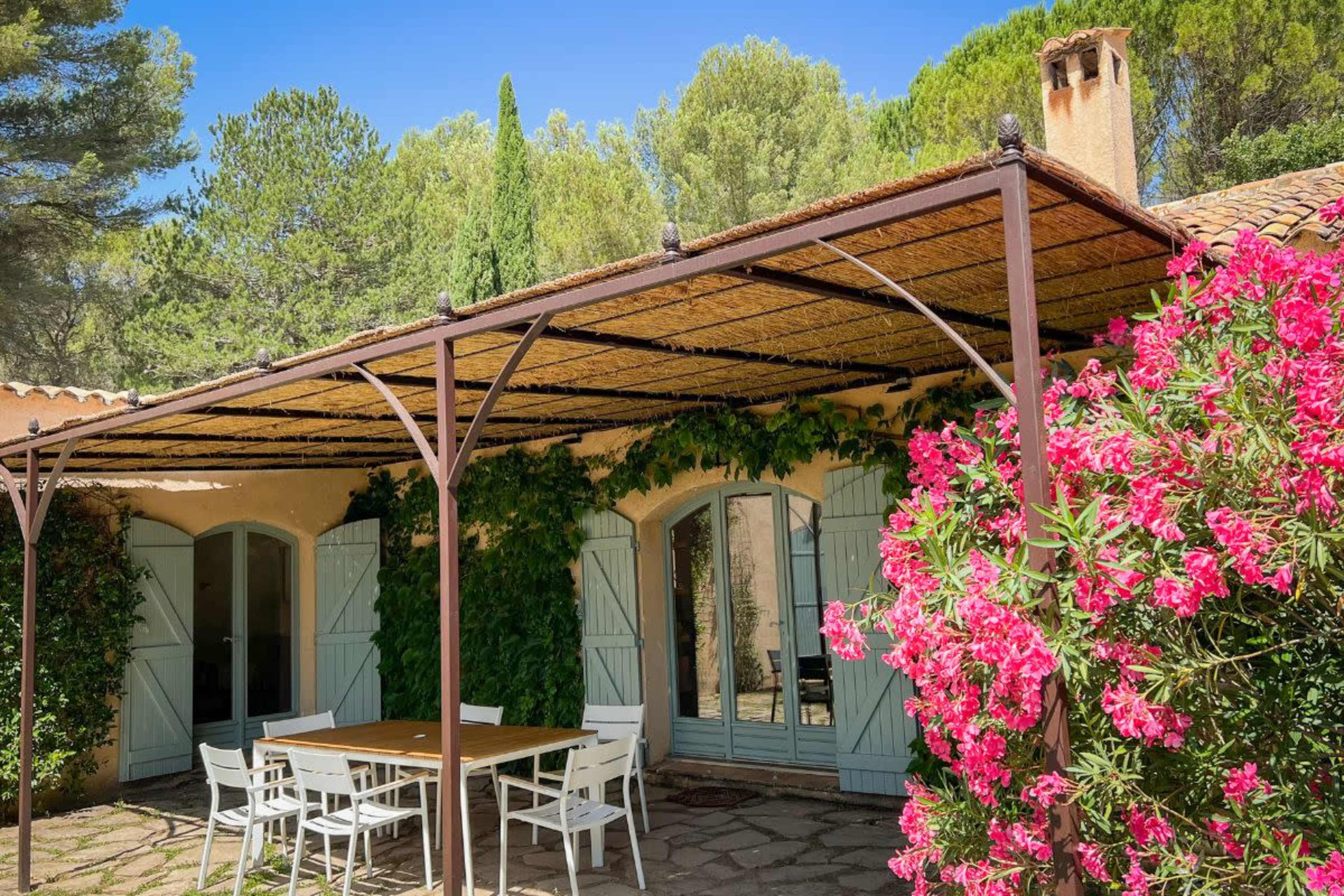 A stone house with light blue shutters is partially shaded by a pergola covered in greenery, surrounded by vibrant pink flowers and trees.