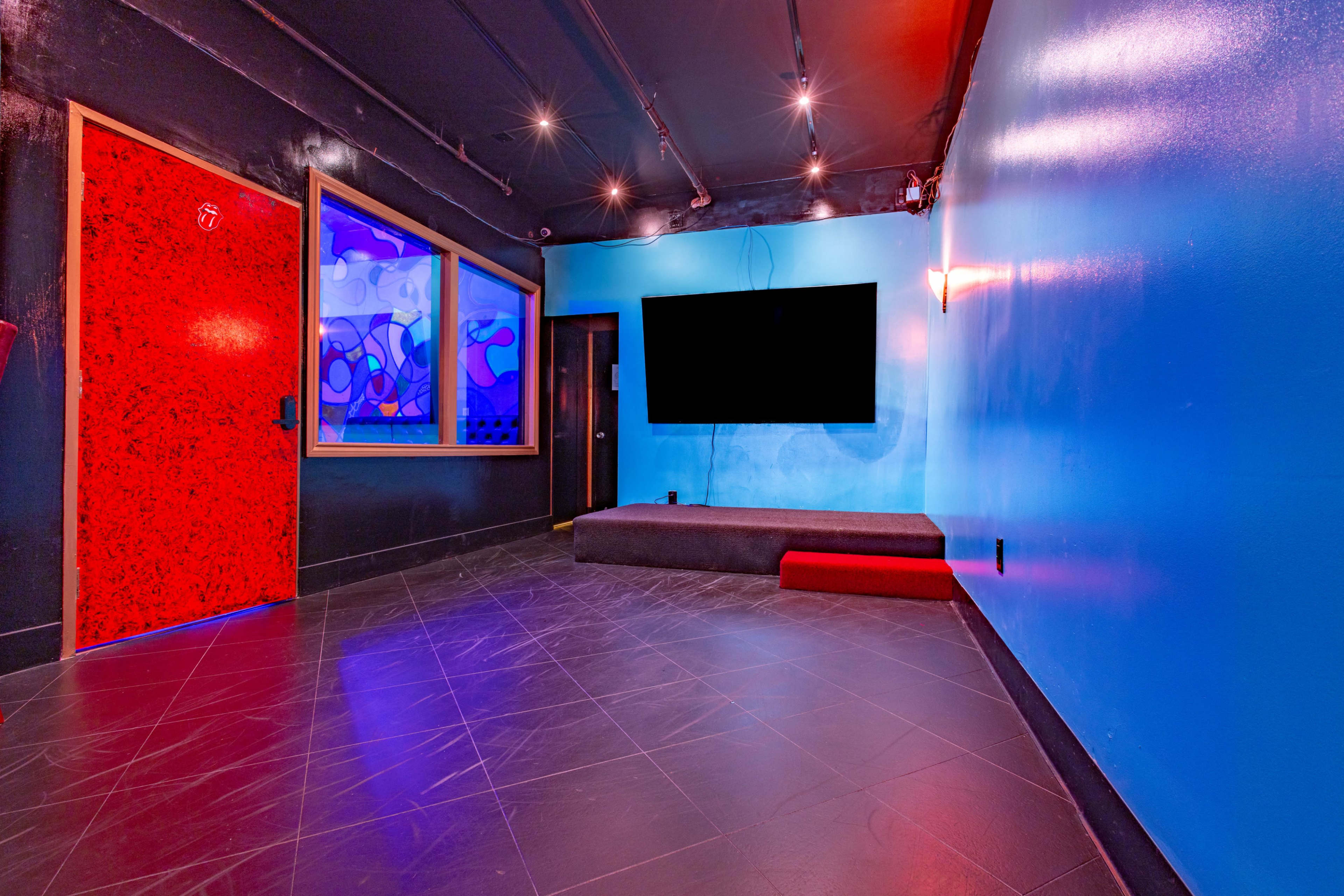 The image shows a dimly lit room with colorful walls, a large television mounted on one wall, and a red door contrasting with the blue surroundings.