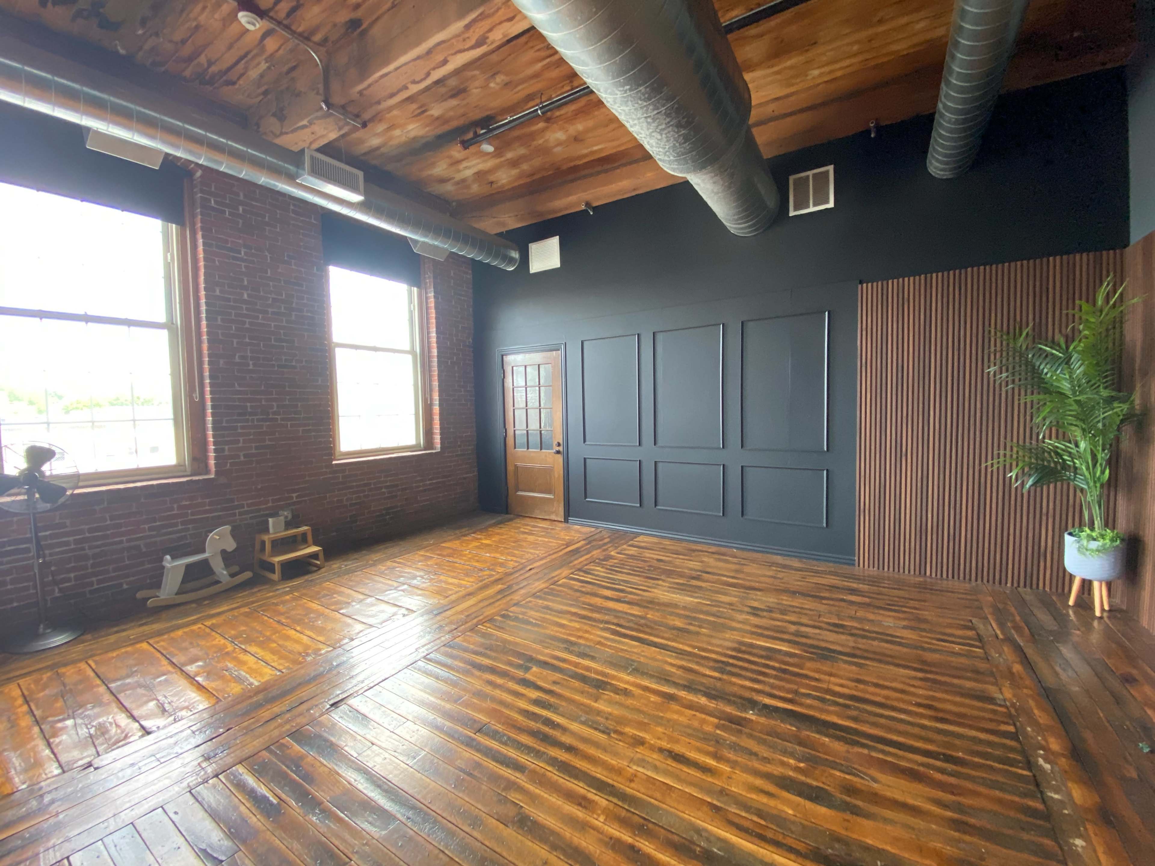 A spacious room with wooden floors, exposed brick walls, and a black accent wall featuring vertical panels, along with a door and large windows allowing natural light.