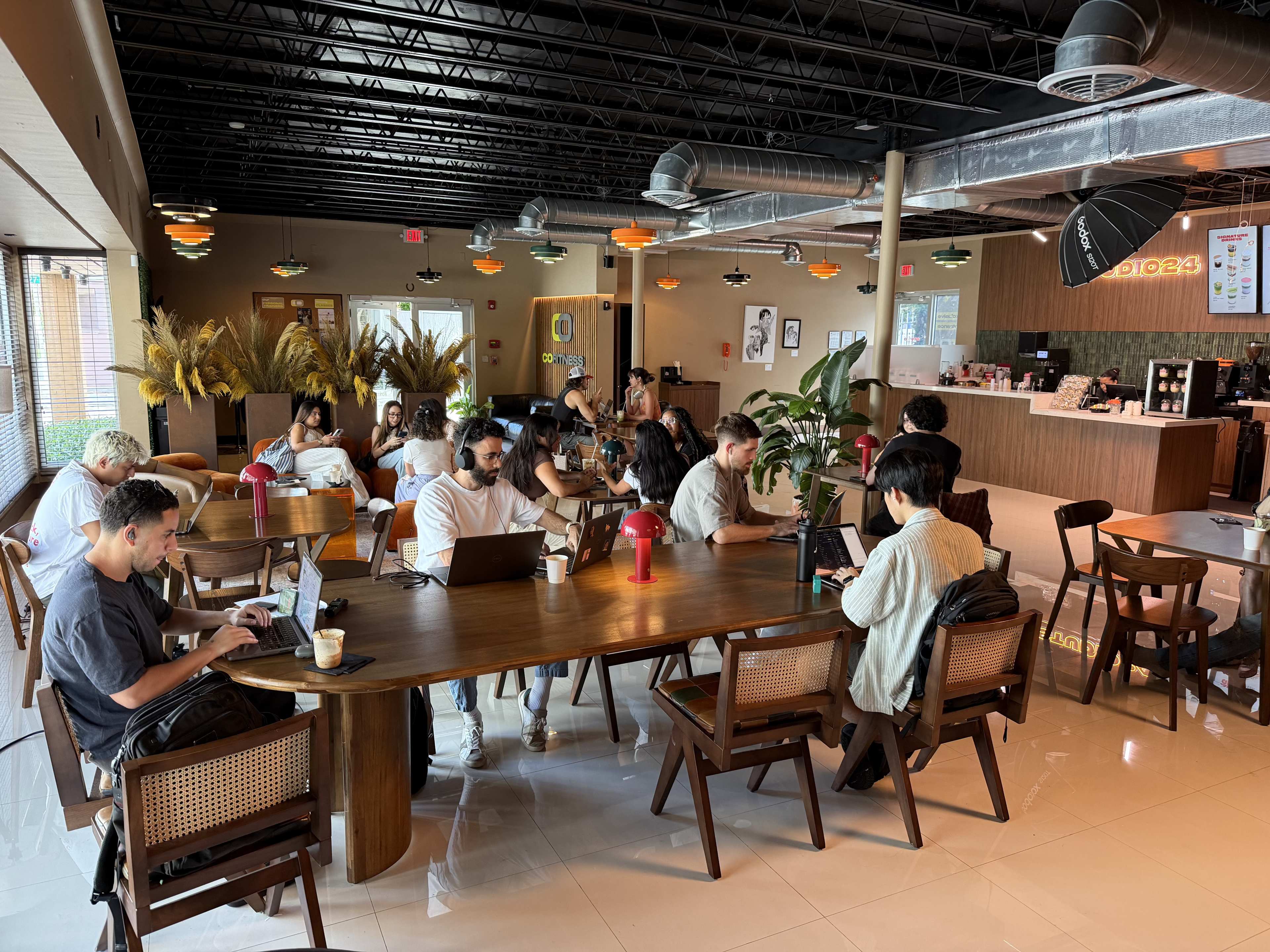 A group of people works at large wooden tables in a modern café, surrounded by potted plants and colorful light fixtures.
