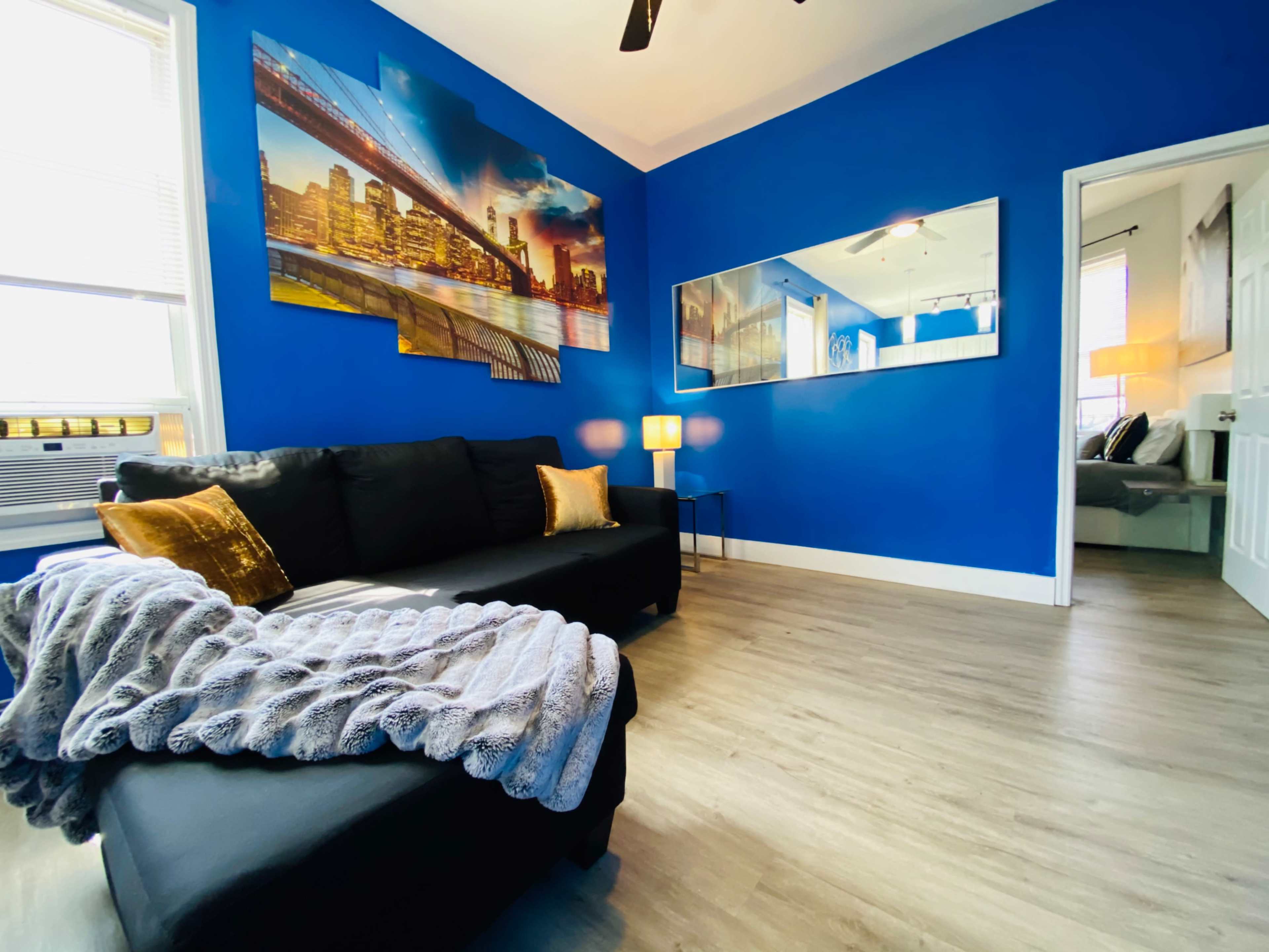 A living room with a blue wall, a black sofa adorned with a gray throw blanket, and a large cityscape painting above it, complemented by two lamps and a doorway leading to another room.