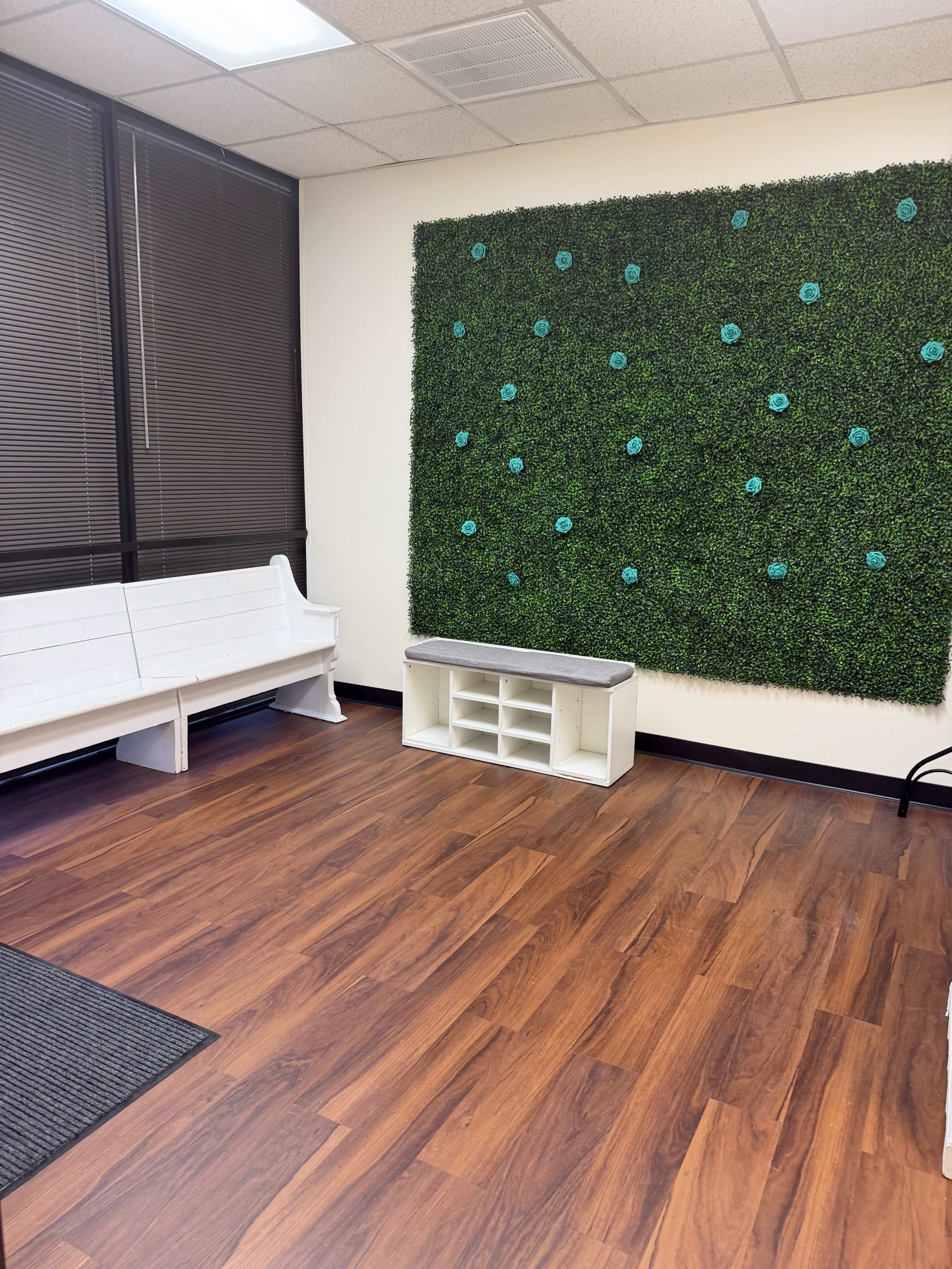 The image shows a room with a wooden floor, a bench on one side, and a green wall decoration featuring artificial flowers.