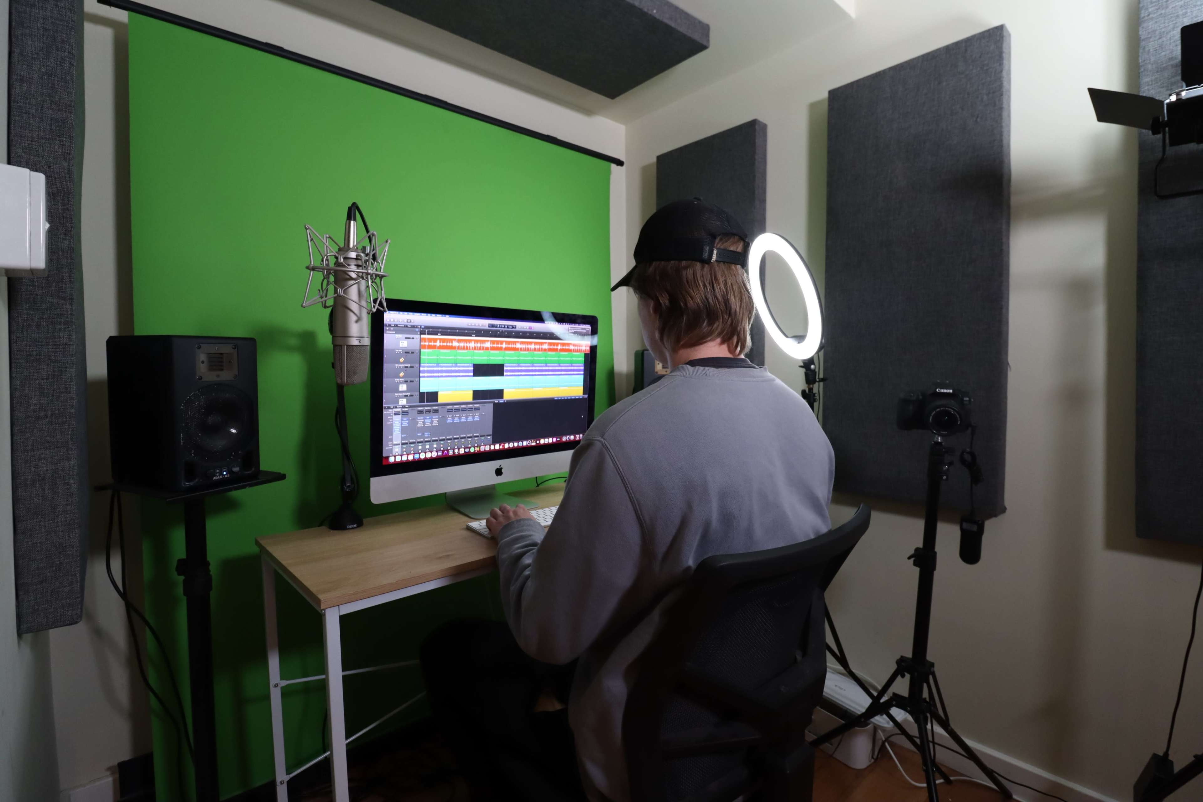 A person sits at a desk in a soundproof studio, working on music production with a computer, microphone, and various recording equipment.