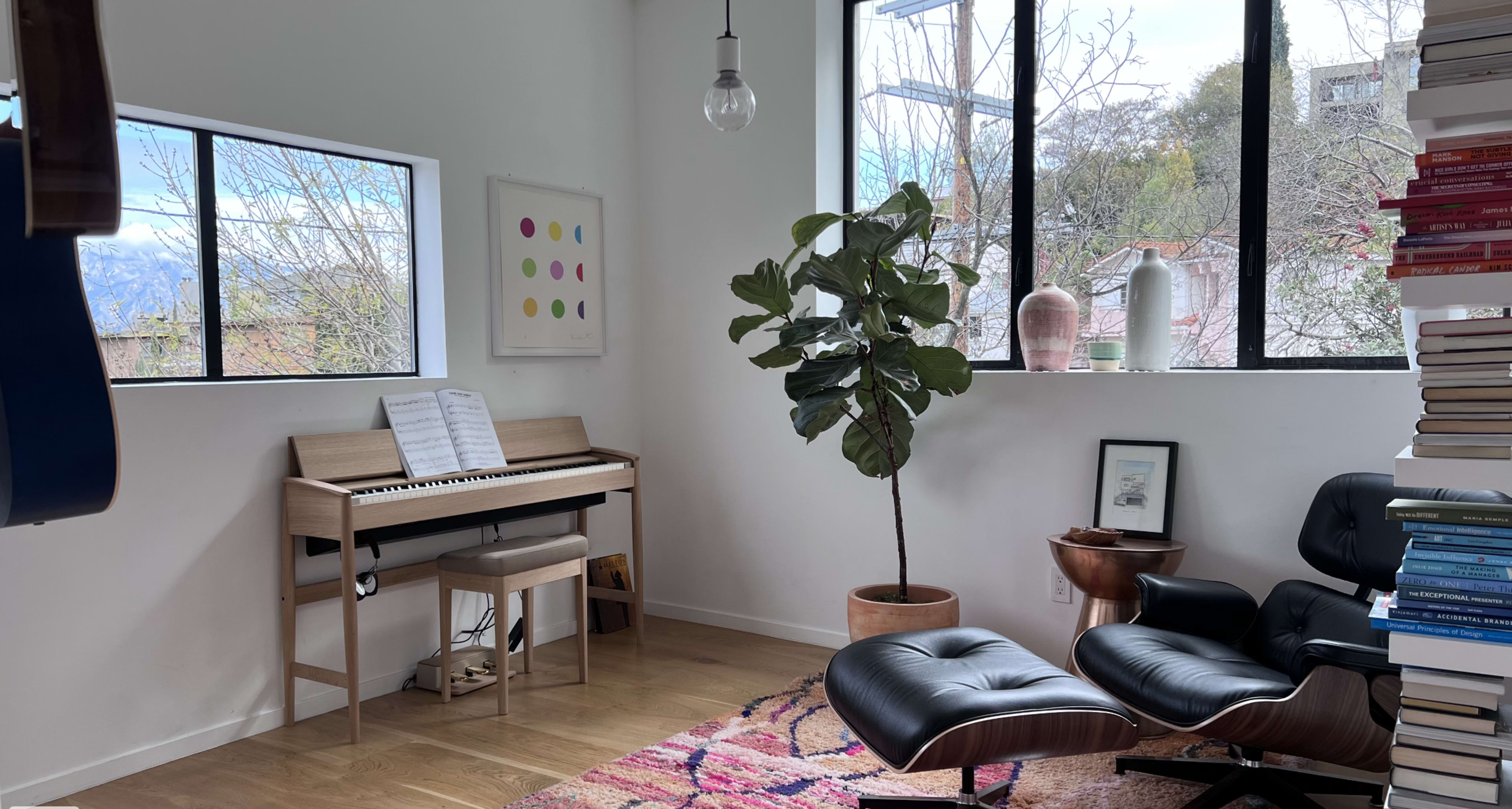 A modern room featuring a piano, a plant in a planter, and bookshelves filled with books, all set against large windows displaying a view of trees.