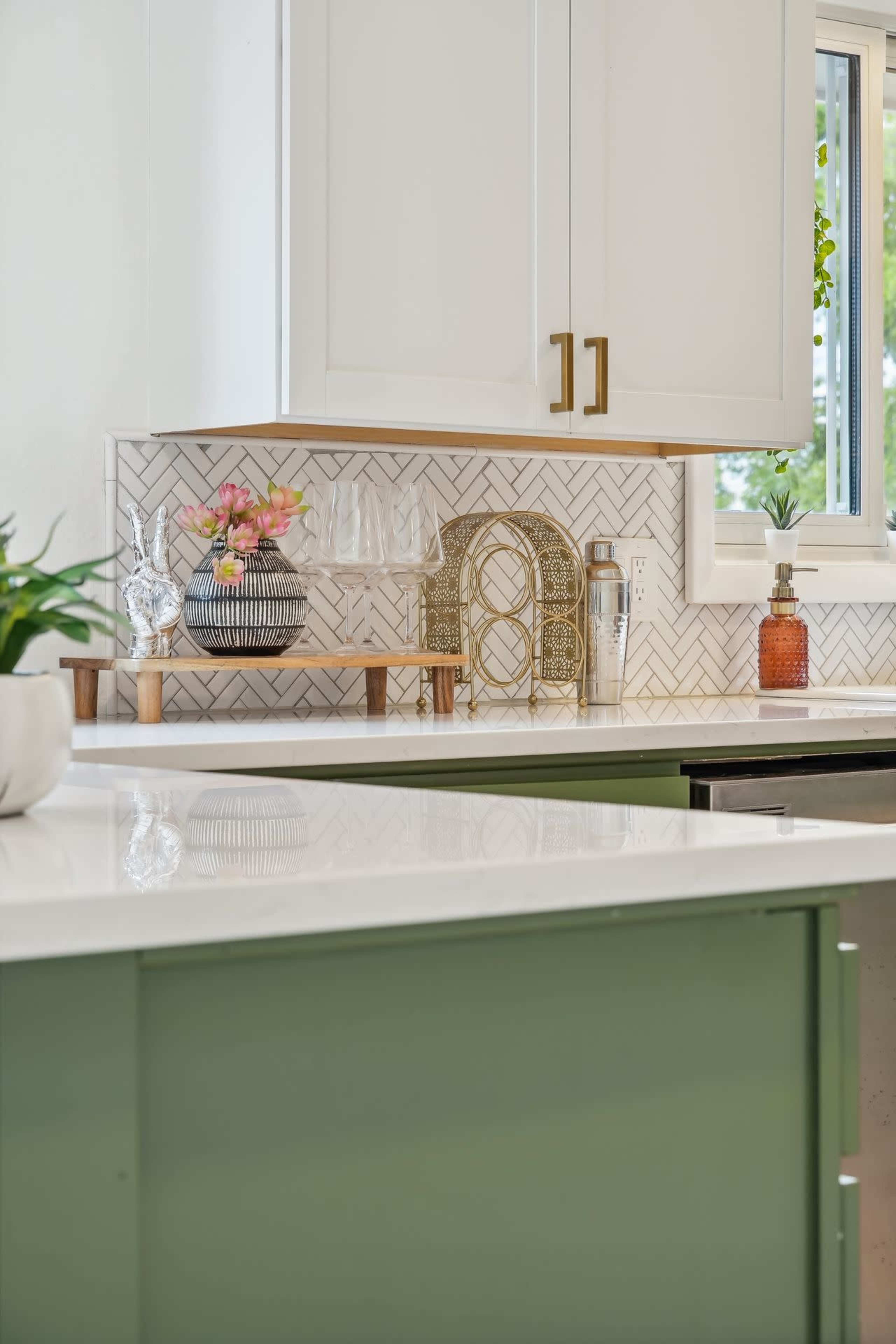 A modern kitchen with white cabinets, a herringbone-patterned backsplash, and a green island countertop featuring decorative items and plants.