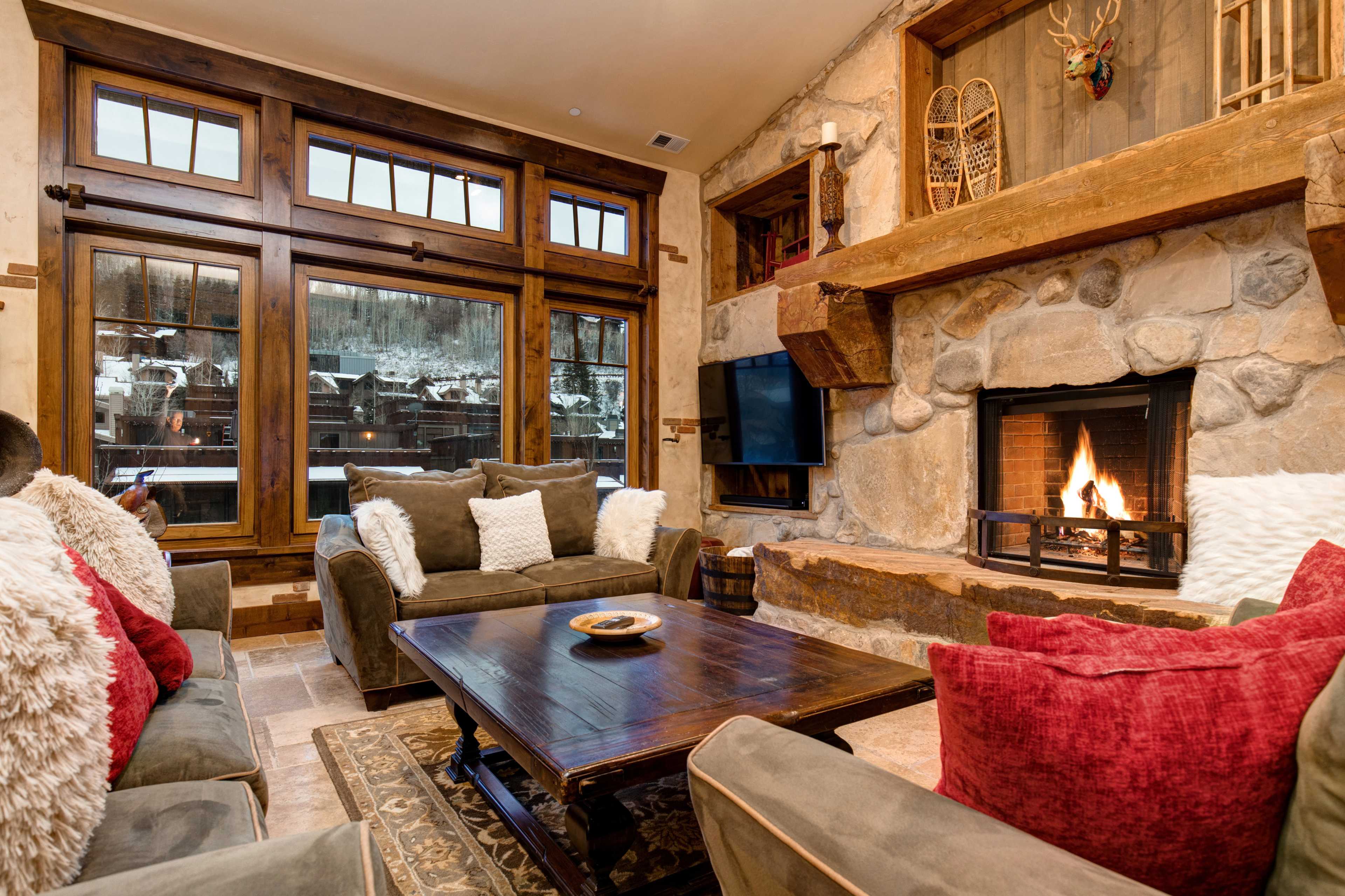 A cozy living room features a stone fireplace, wooden accents, and large windows overlooking a snowy landscape.