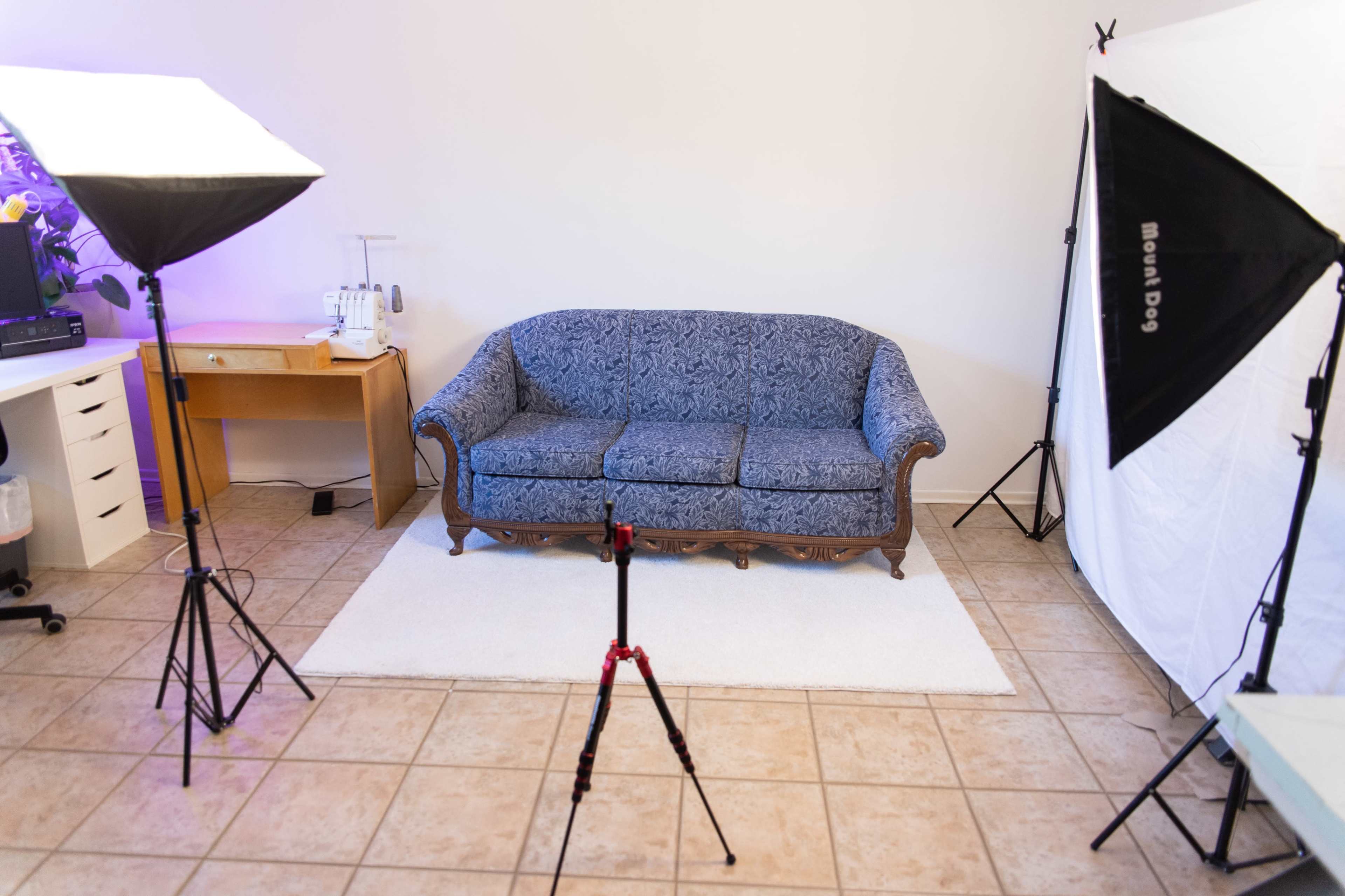 A blue patterned sofa sits on a white rug in an indoor space, surrounded by studio lighting equipment and a sewing machine on a nearby table.
