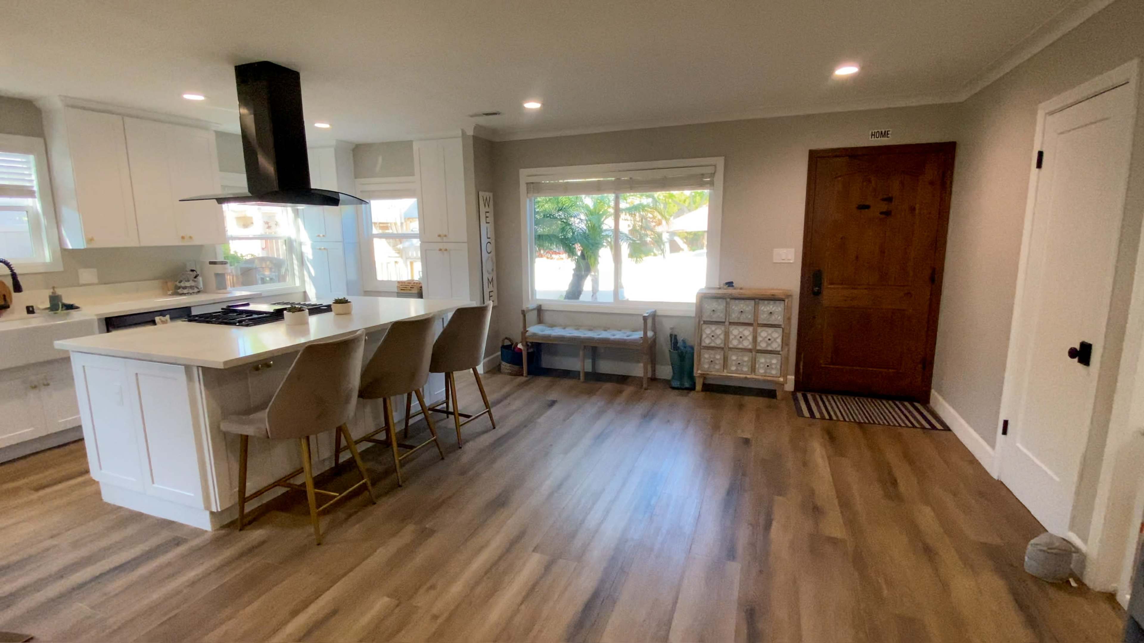 The image shows a modern kitchen and entryway with a breakfast bar, a front door, and a large window letting in natural light.