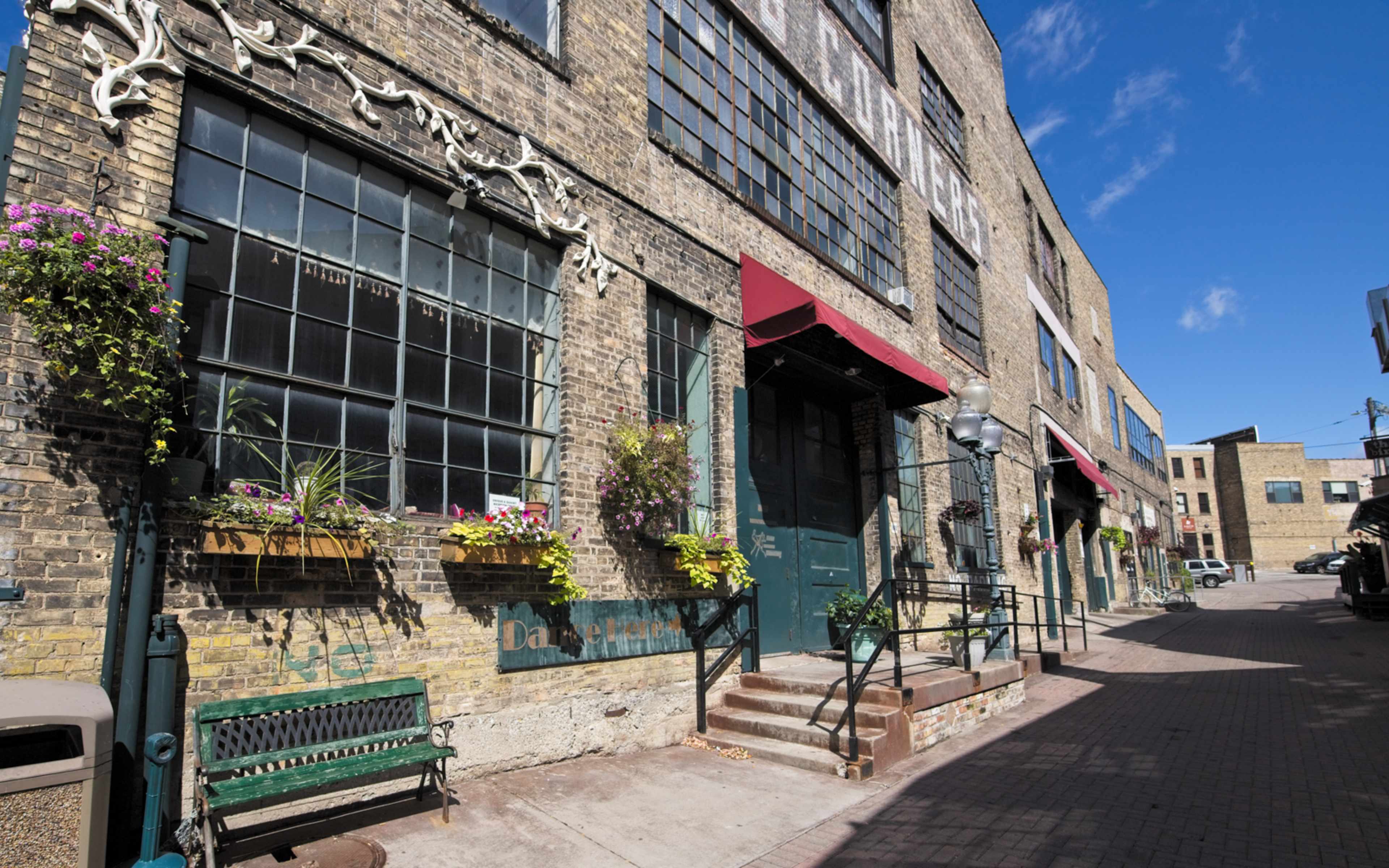 A row of brick buildings featuring large windows and awnings, with planters filled with flowers along the lower level and a concrete walkway in front.