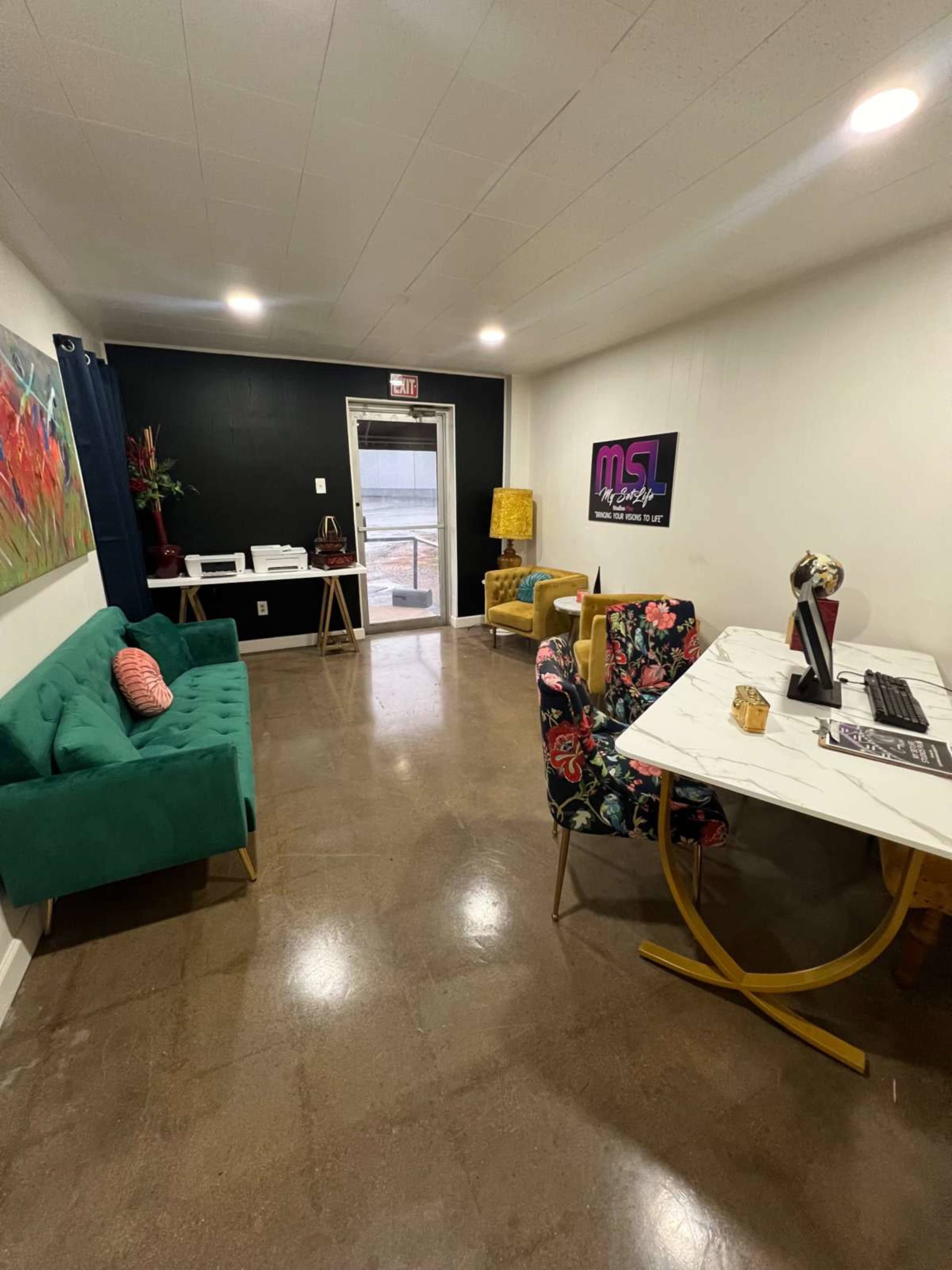 The image shows a modern office space with a green sofa, a floral-patterned chair, a white desk with a computer, and a large window providing natural light.