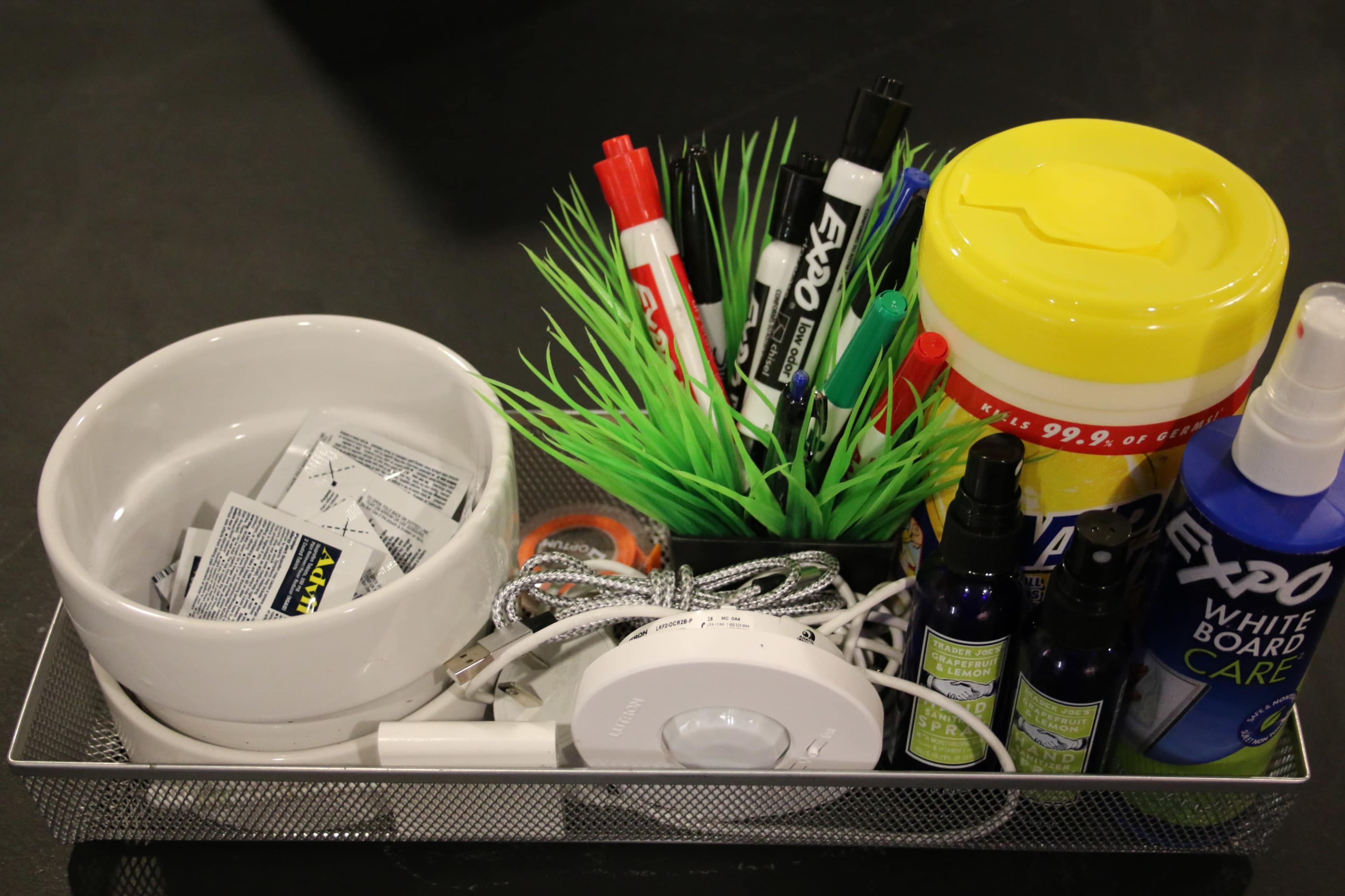 A metal tray contains a white bowl with packets, colorful markers, a yellow container, and various bottles, alongside a small decorative plant.
