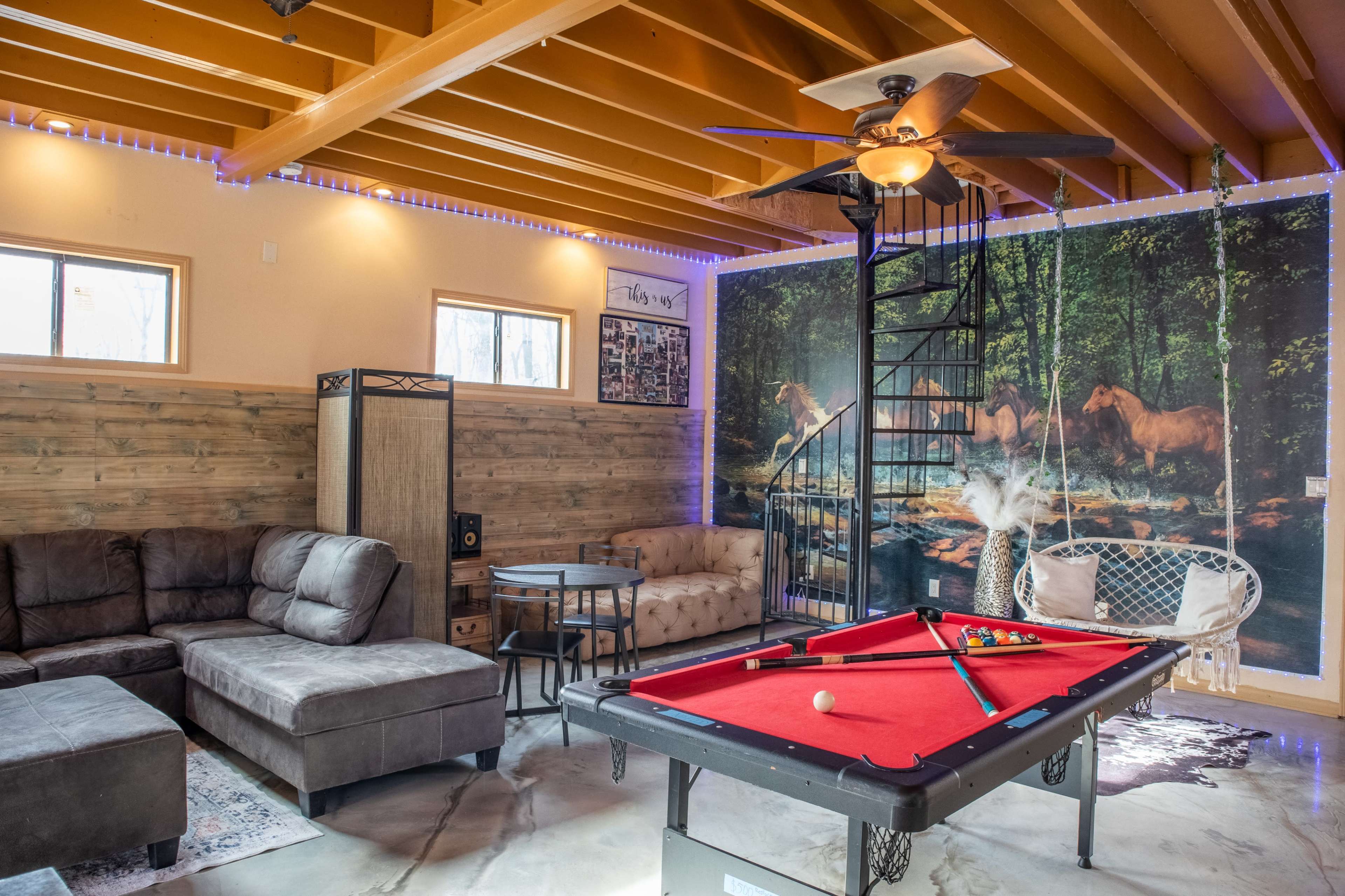 The image shows a recreational room featuring a red pool table, a large sectional sofa, and a spiral staircase beside a mural of horses in a forest.