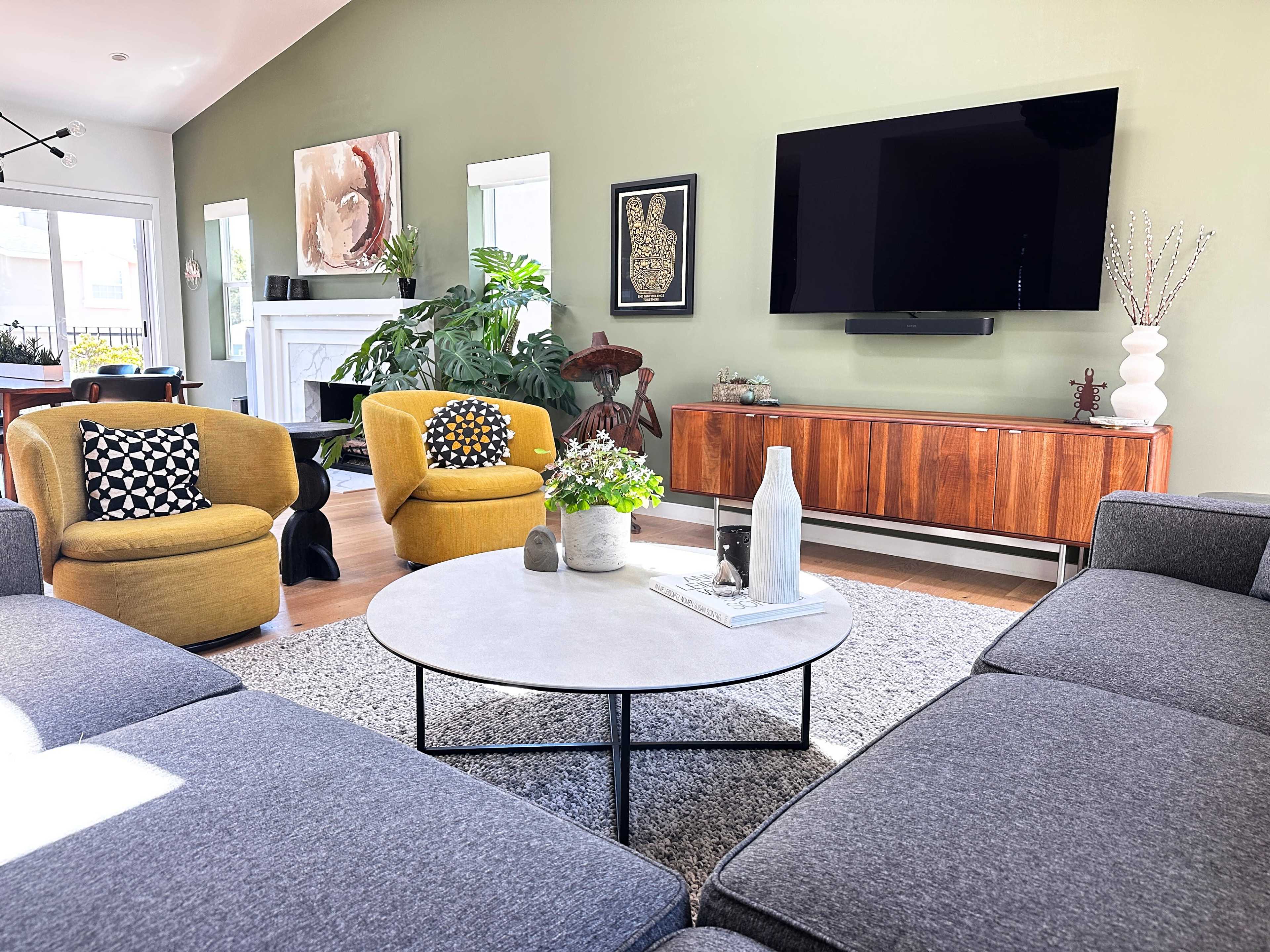 A modern living room features a gray sectional sofa, a round coffee table, and two yellow armchairs with patterned cushions, complemented by green walls and a television mounted on the wall.