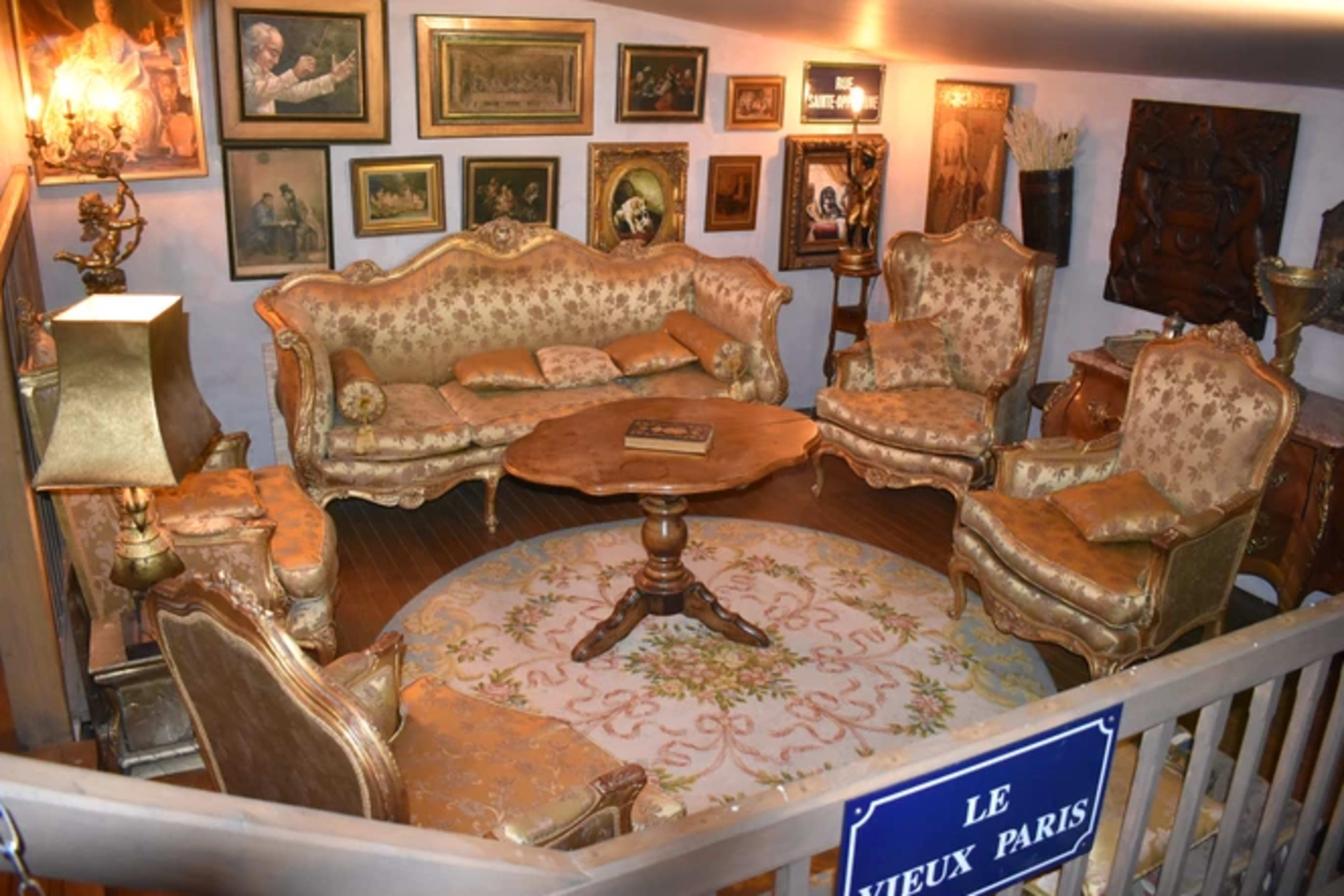 A vintage-style living room furnished with ornate seating and a round wooden table, surrounded by framed artwork on the walls.
