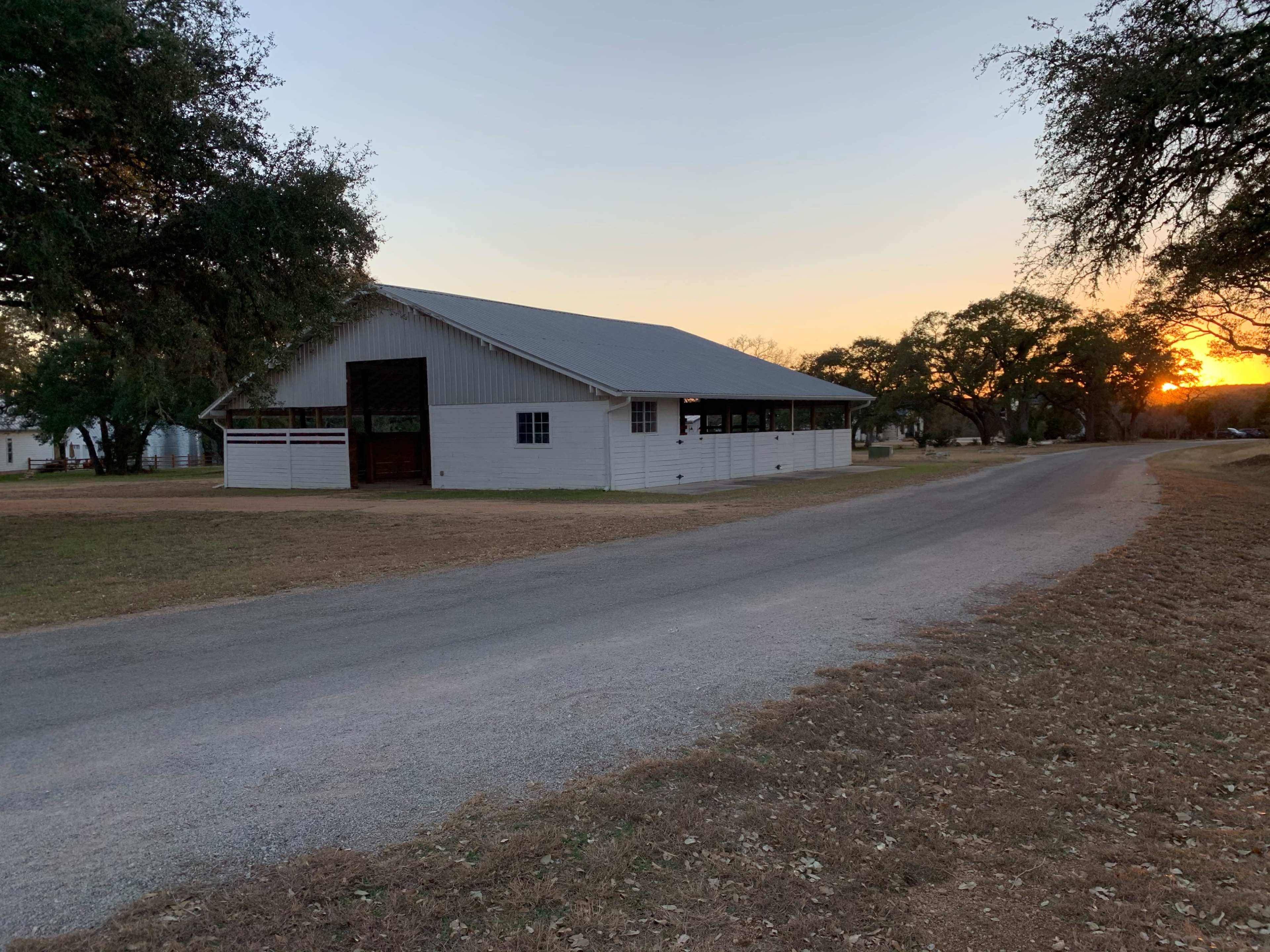 A white building with a metal roof stands beside a gravel road at sunset, surrounded by trees.
