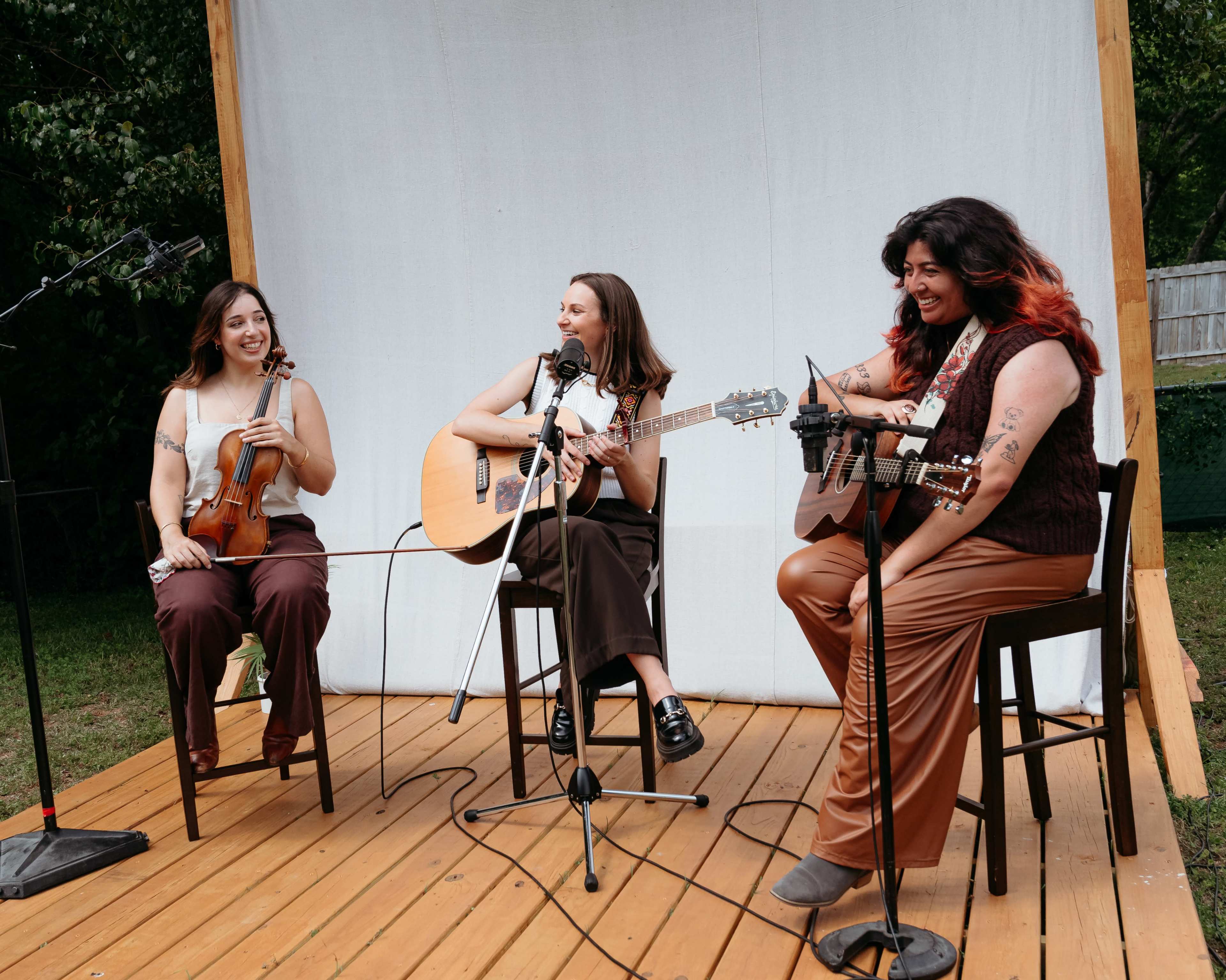 Three musicians are performing on a wooden stage, playing a violin and two acoustic guitars, under a white backdrop in an outdoor setting.