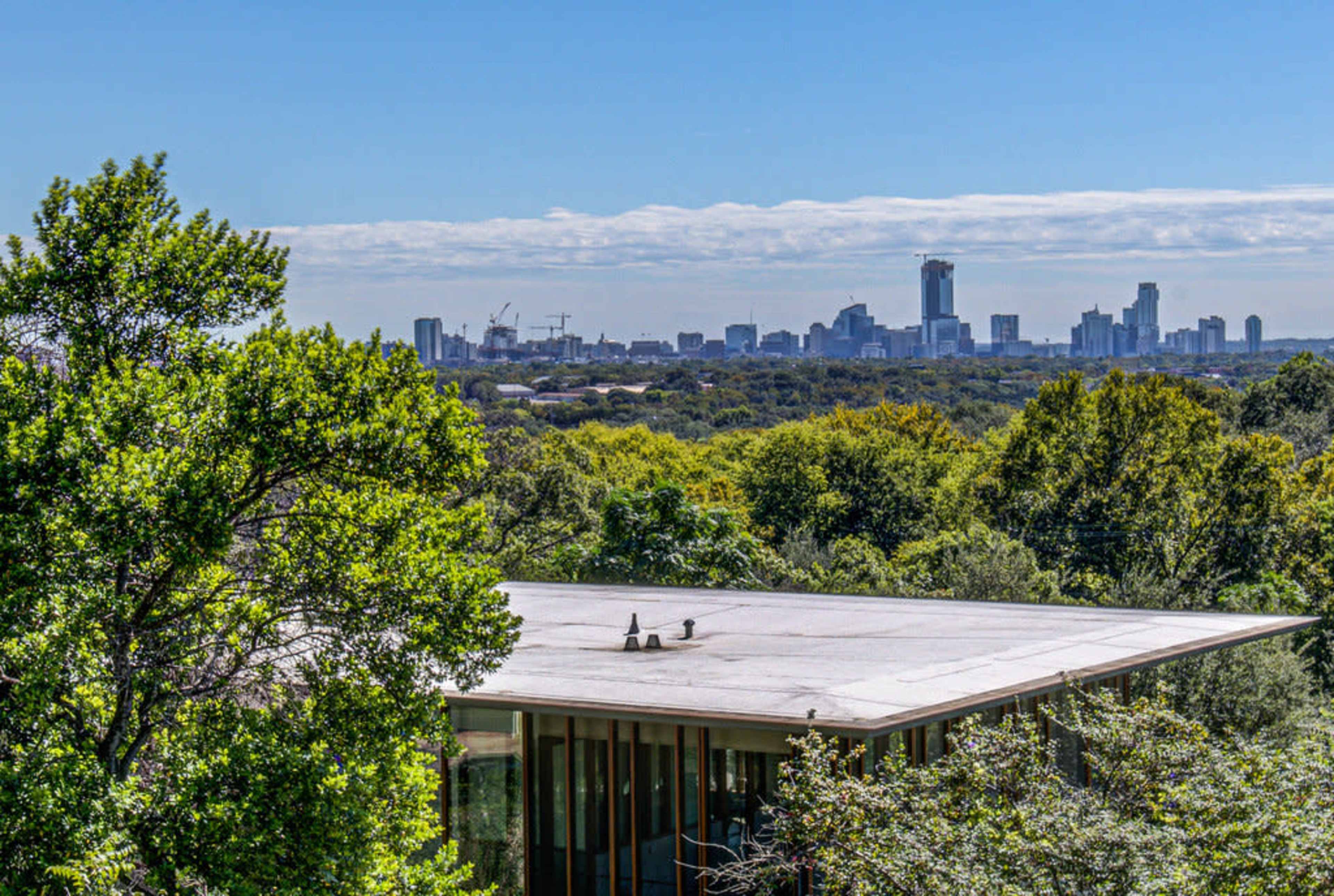 The image shows a modern building roof in the foreground with a skyline of a city visible in the background, surrounded by green trees and open landscape.