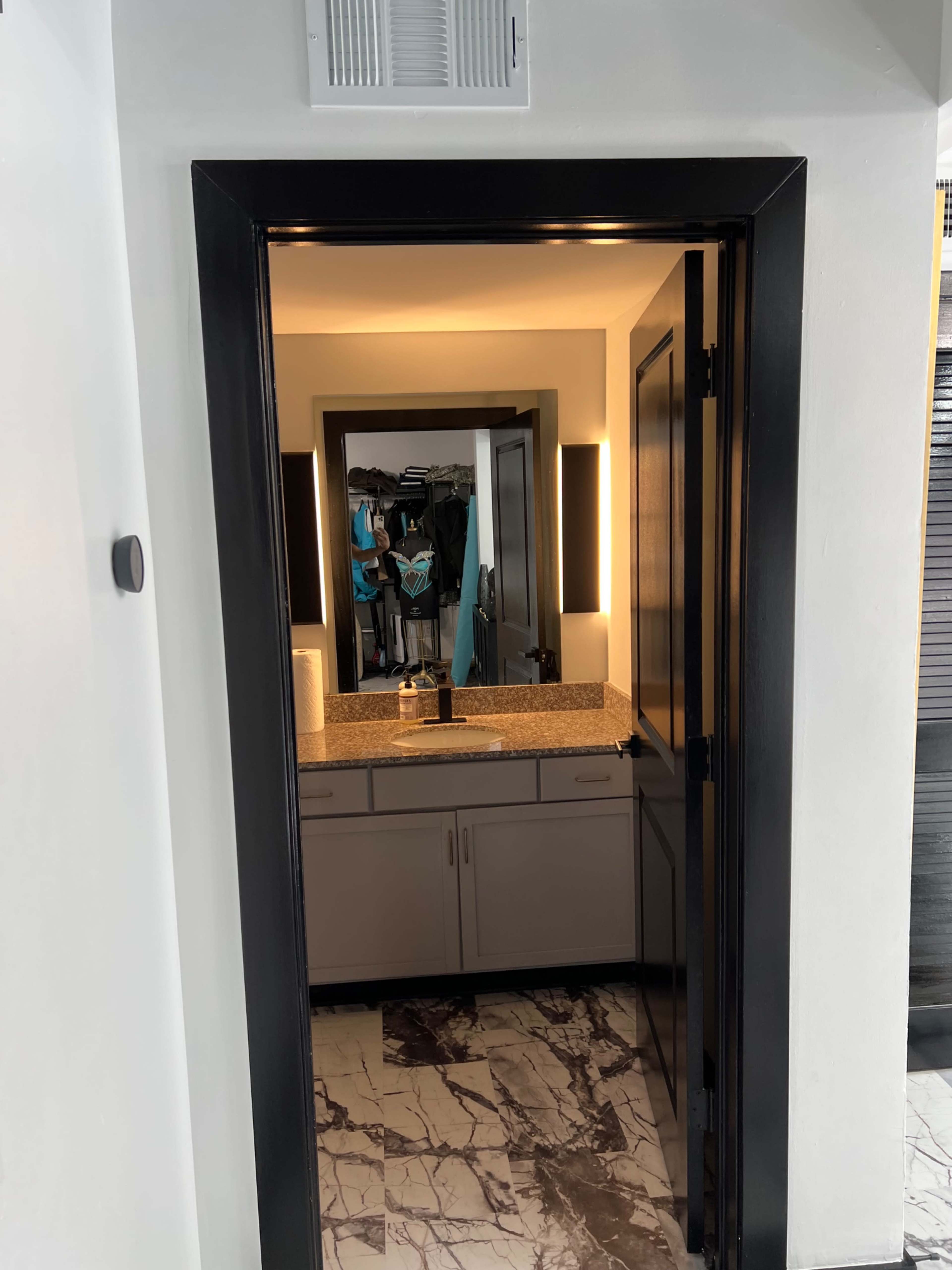 The image shows a doorway leading into a bathroom with a double-sink vanity and a large mirror, surrounded by lighted sconces.
