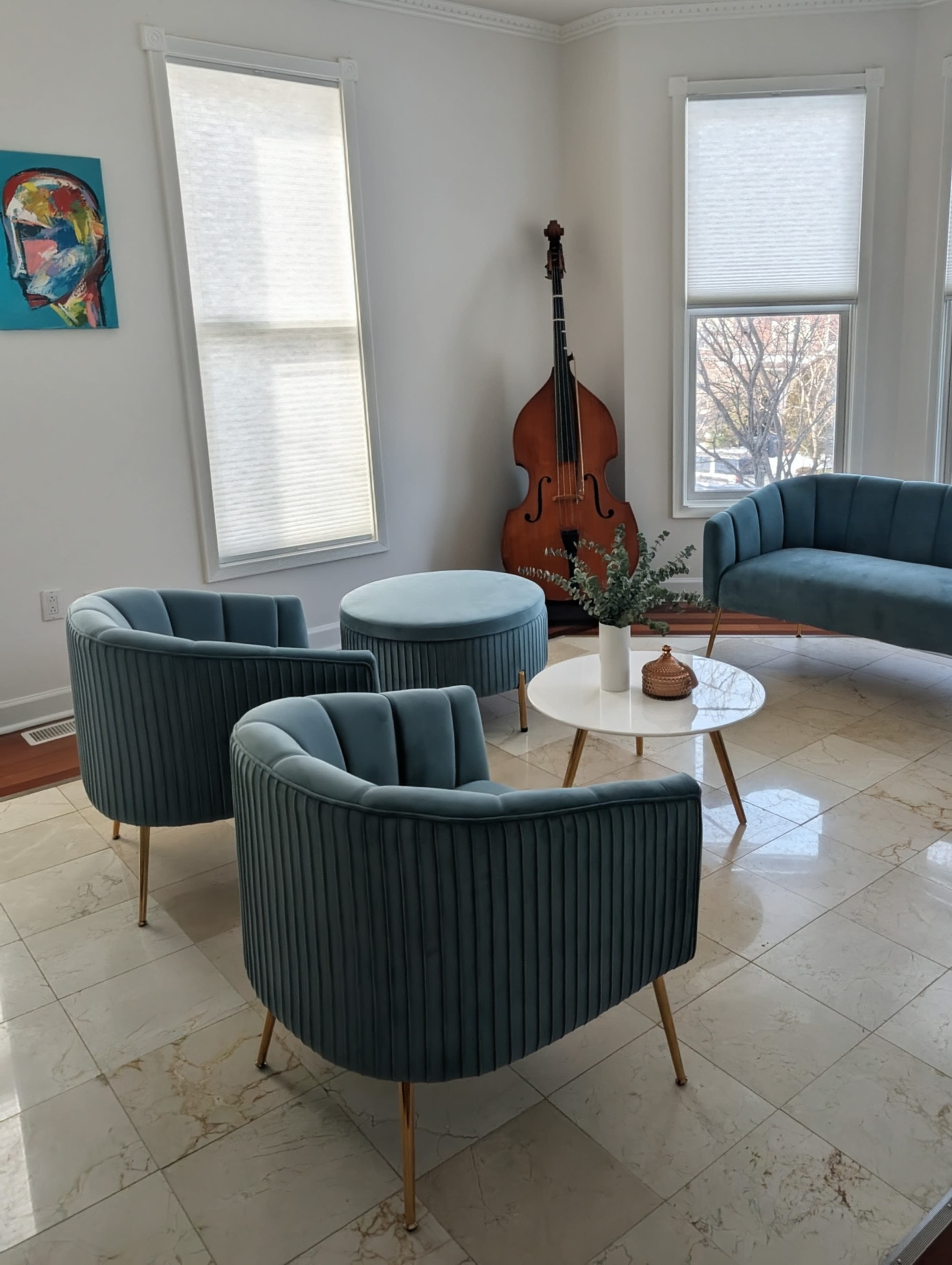 The image shows a sunny interior with two light-blue upholstered chairs, a small round table, a white coffee table, and a double bass positioned near the window.