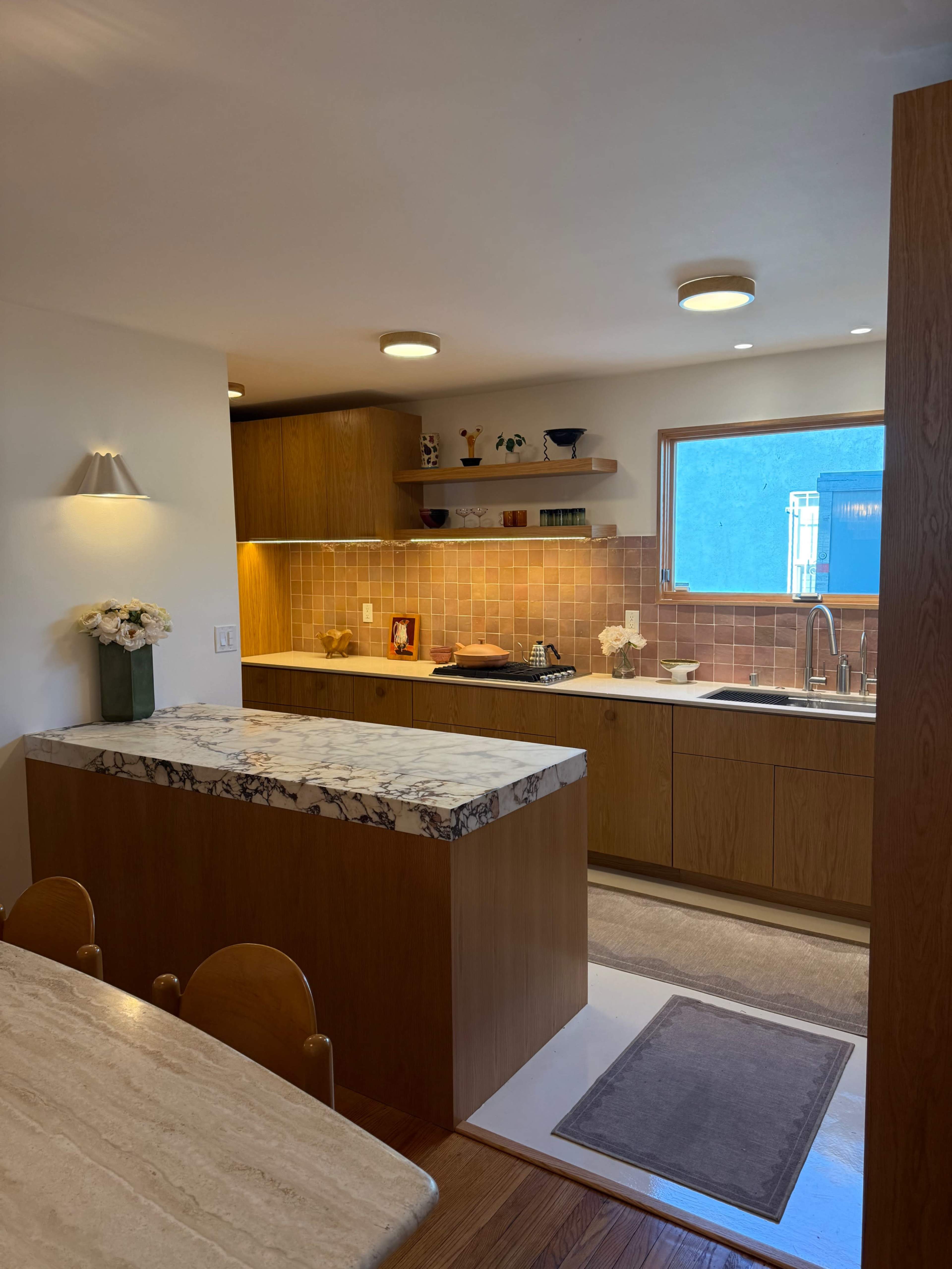 The image shows a modern kitchen featuring wooden cabinets, a marble countertop, and a window providing natural light.