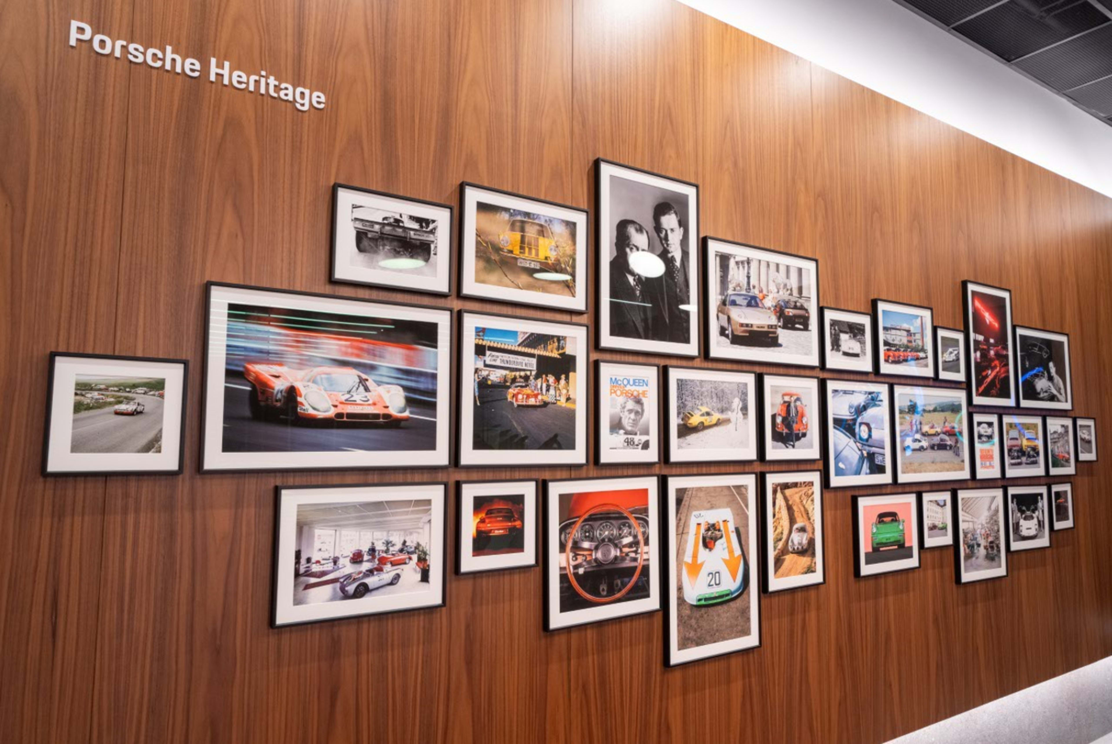 The image features a wooden wall displaying a collage of framed photographs and memorabilia related to Porsche, showcasing various cars, racing events, and historic moments.