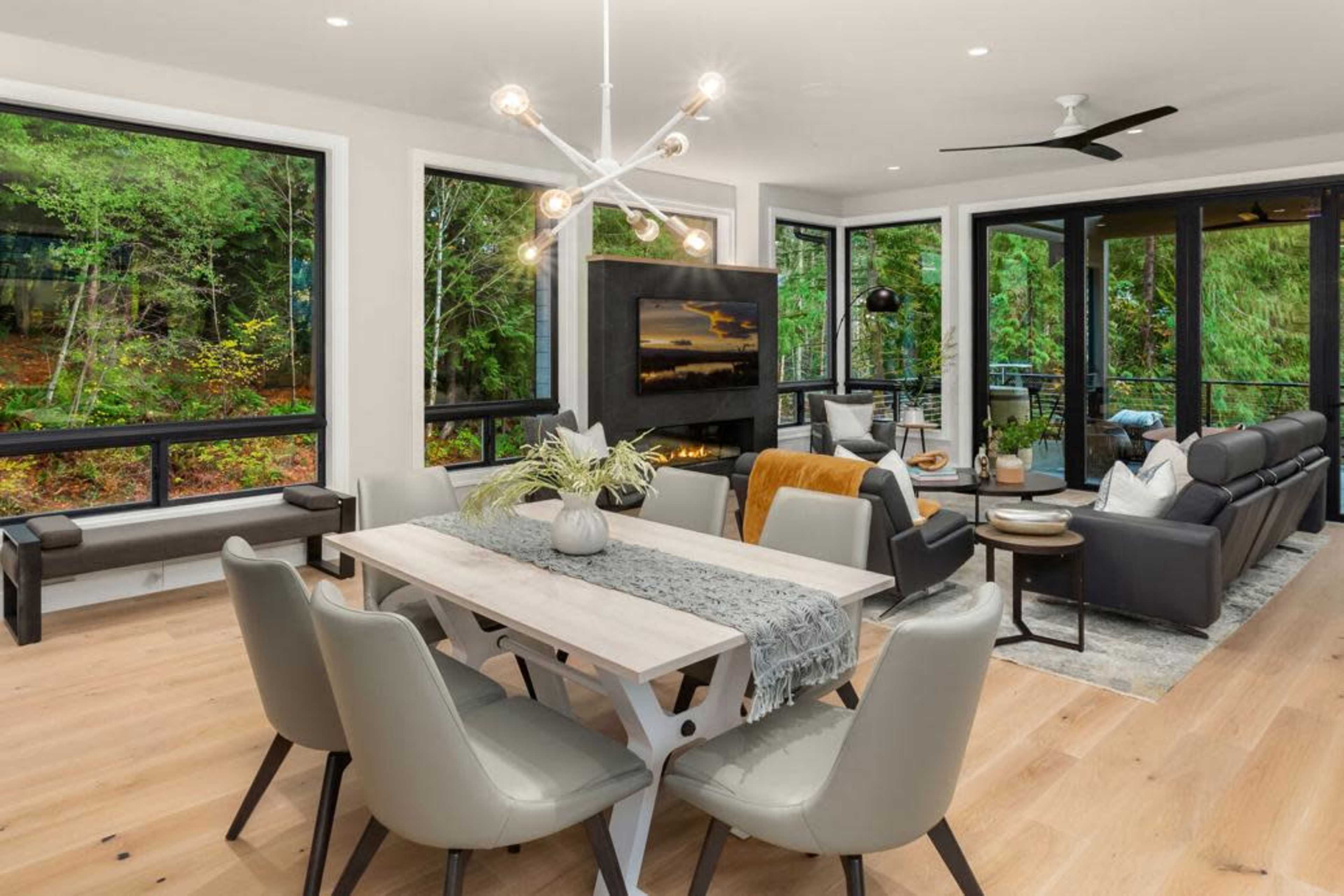 A modern living and dining area features a dining table with chairs, a seating arrangement around a fireplace, and large windows overlooking greenery.