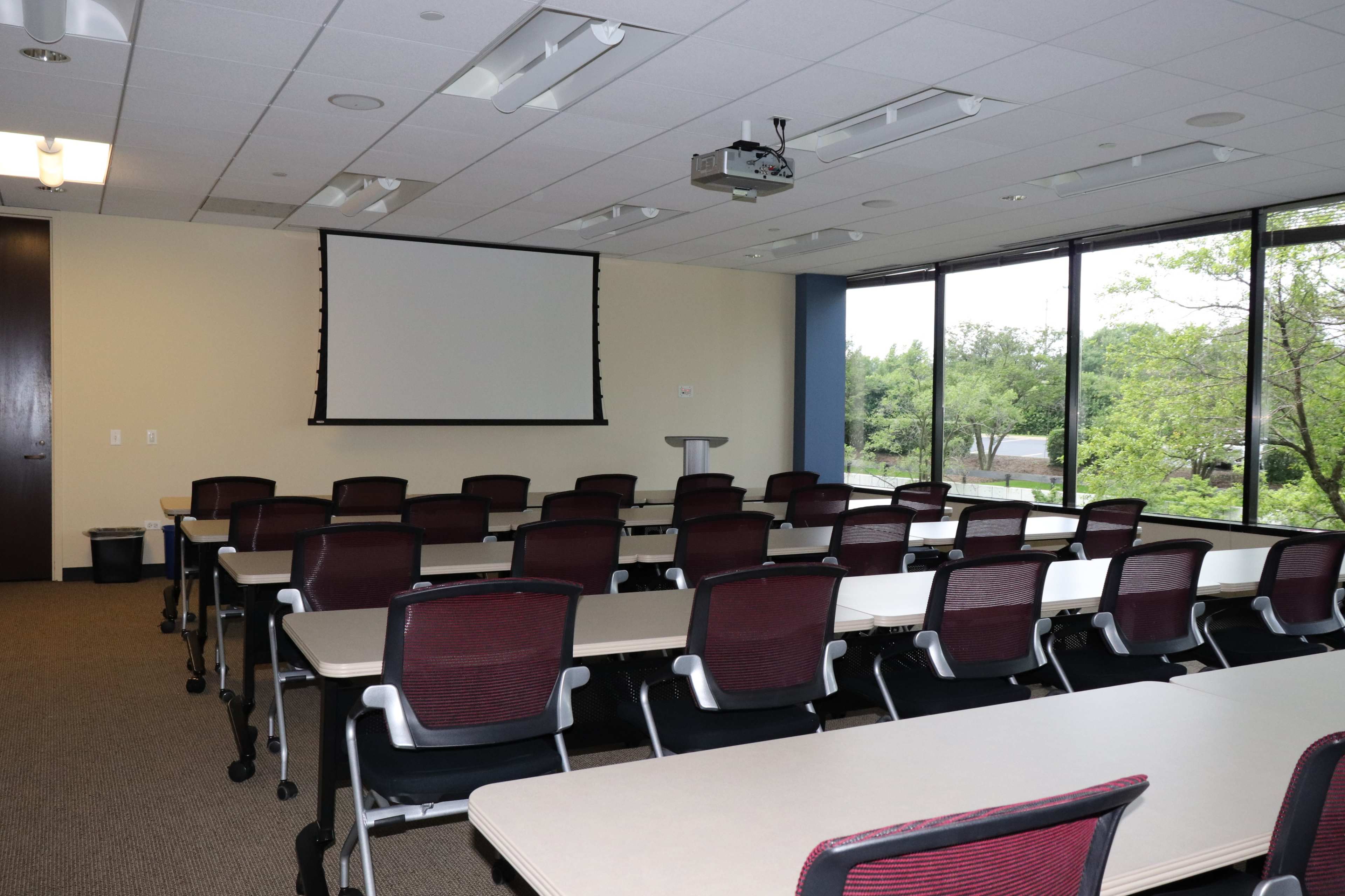 A conference room with multiple tables and chairs arranged for a meeting, featuring a projector screen and large windows providing natural light.