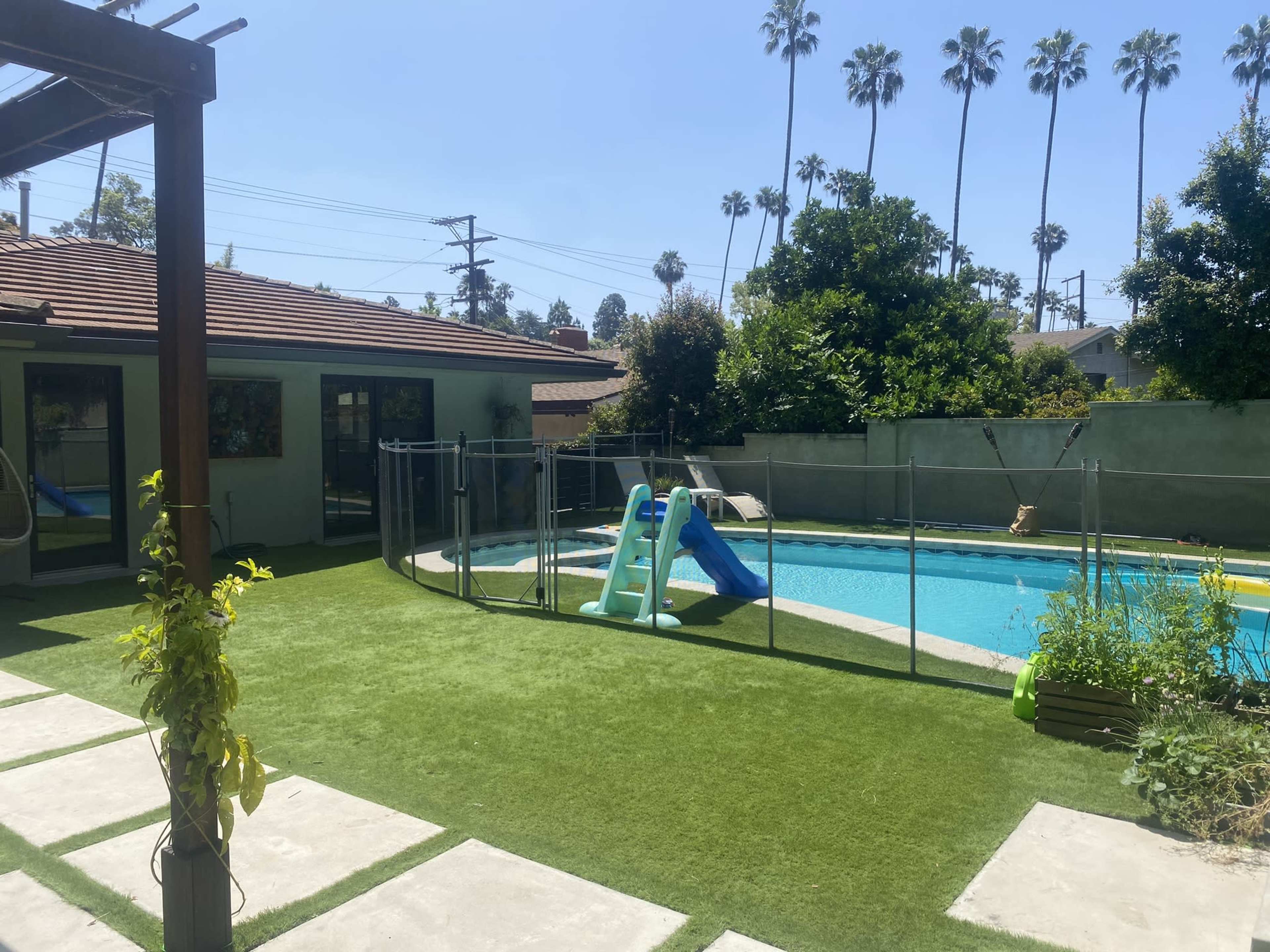 A backyard with a swimming pool, a slide, a lush lawn, and palm trees in the background.